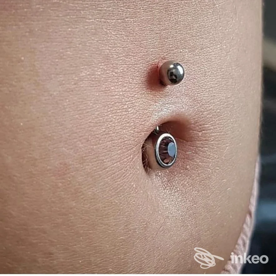 Piercing visibile sull’ombelico: due piercing metallici, uno piccolo in alto e uno anello sottostante, su pelle neutra. Studio Mercante d'Arte.