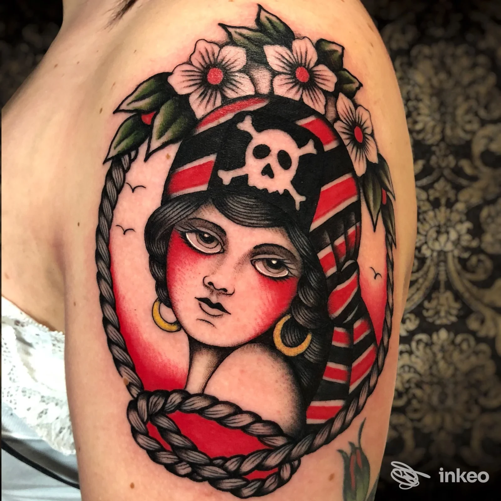 Tattoo a colori su pelle reale nella parte superiore del braccio: ritratto di una donna con cappello contenente un teschio, cornice di fiori e una corda intrecciata; colori vividi e linee nette.