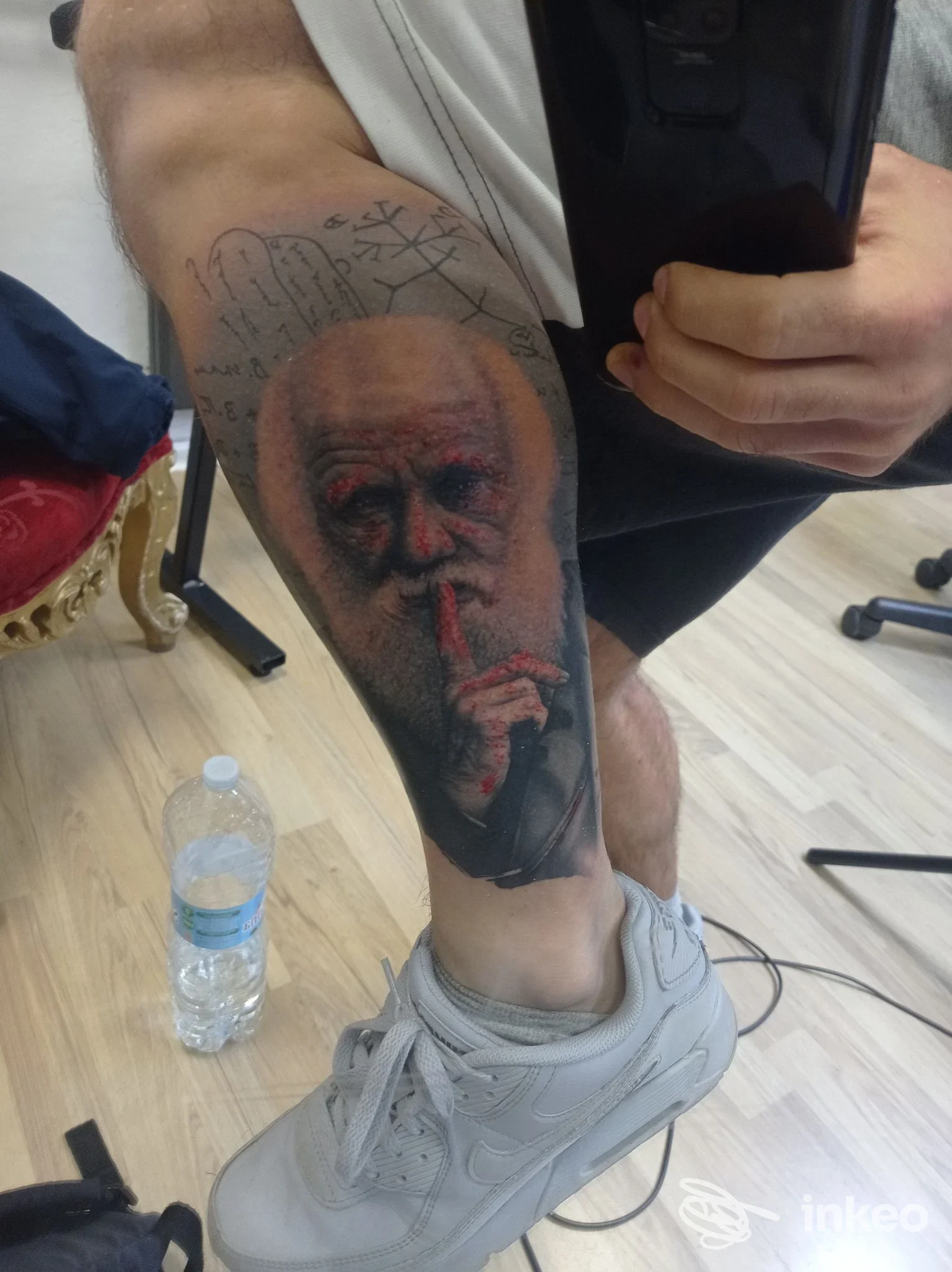 Tattoo realistico su gamba di un uomo anziano, primo piano della parte inferiore della gamba con l'immagine del volto visibile; nessun volto intero è mostrato.