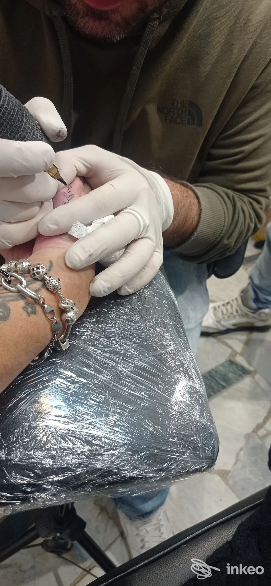 Procedura di tatuaggio in corso su pelle umana: mani dell’artista con guanti bianchi maneggiano l’ago, versione parziale del viso dell’artista visibile, braccio e polso con catena e bracciali, superficie protetta in lamina metallica.