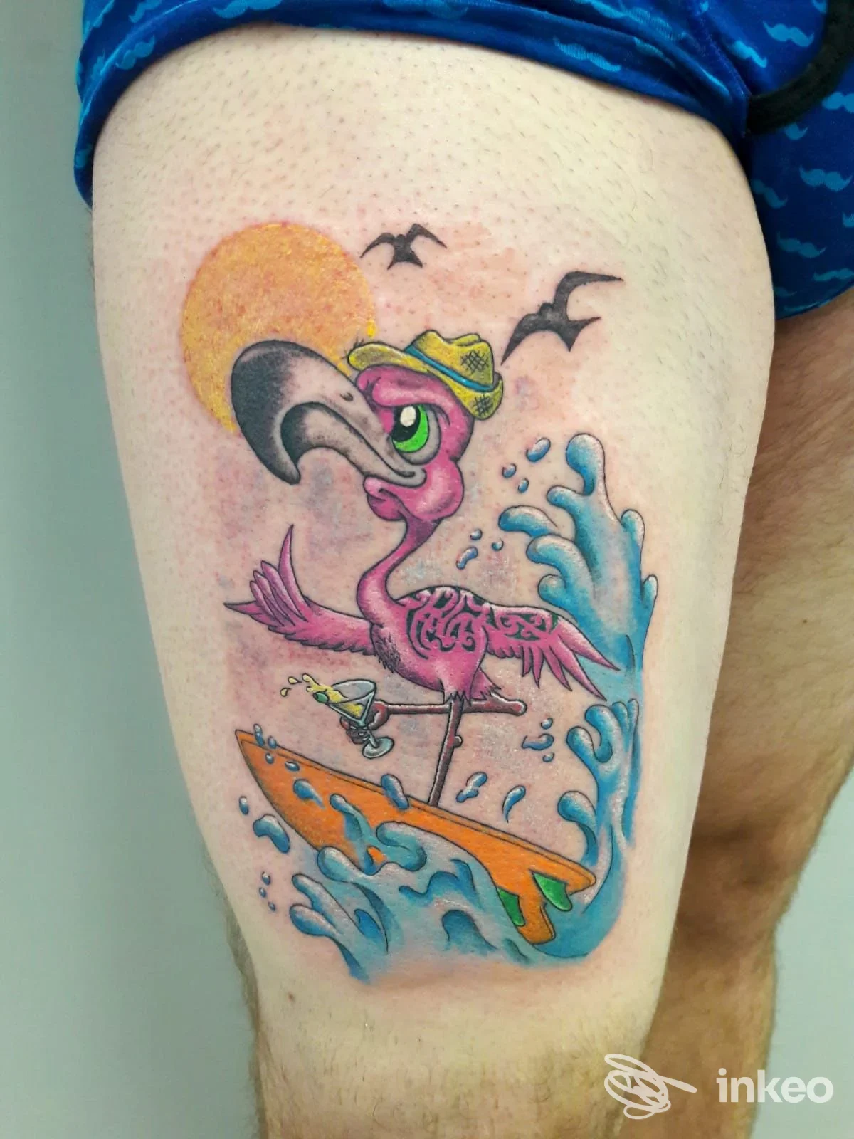 Tattoo colorato di un fenicottero rosa con tavola da surf, sole e onde, sulla pelle della parte superiore della gamba, senza volto visibile.