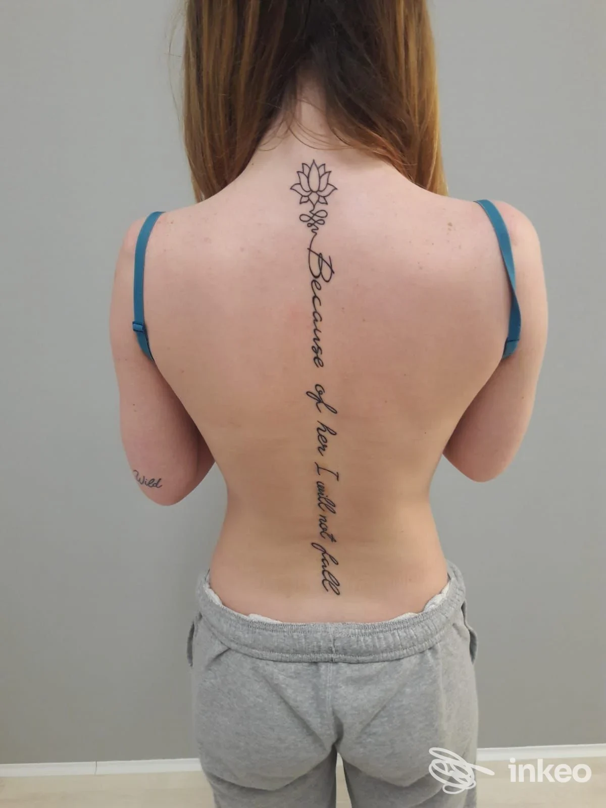 Tattoo verticale lungo la schiena con fioritura a lotus e testo corsivo centrale, visibile su una persona vista di schiena in studio.