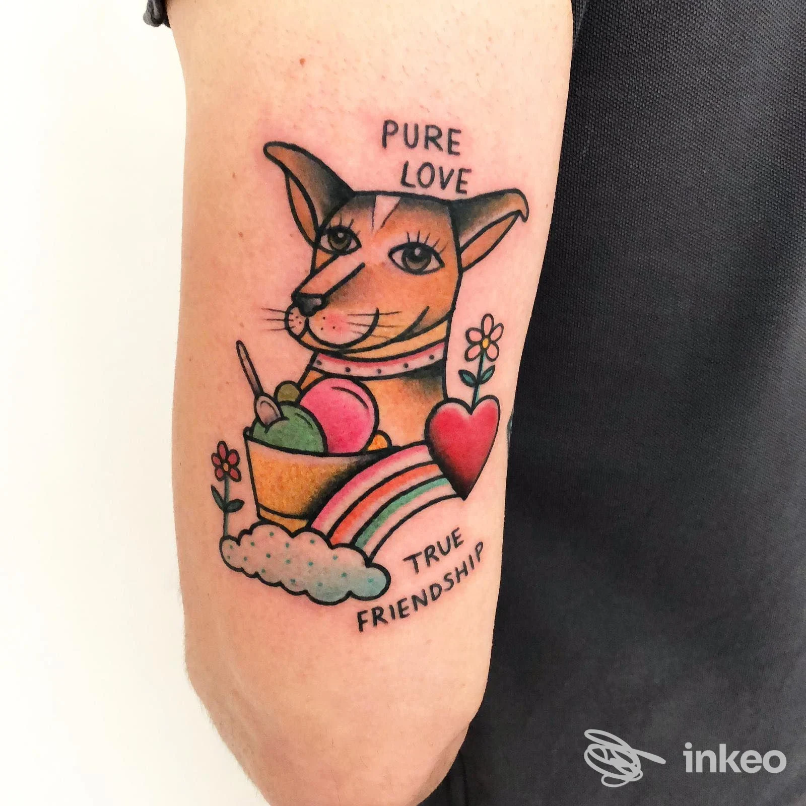 Tatuaggio sull'avambraccio di un gatto stilizzato con fiori, cuore e arcobaleno; testo PURE LOVE in alto e TRUE FRIENDSHIP in basso, colori vivaci.