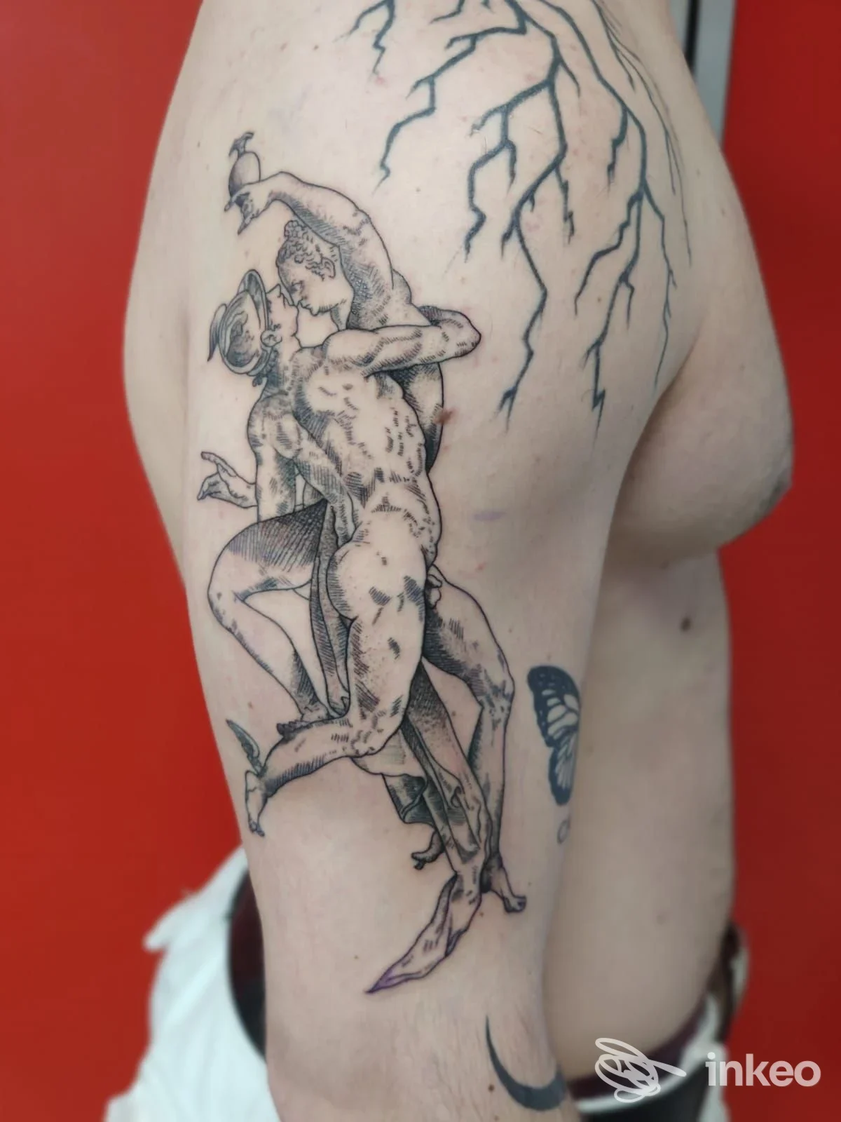 Tattoo in stile scultoreo su braccio sinistro: due figure nude intrecciate, tratteggiate in bianco e nero su pelle chiara, sfondo rosso.