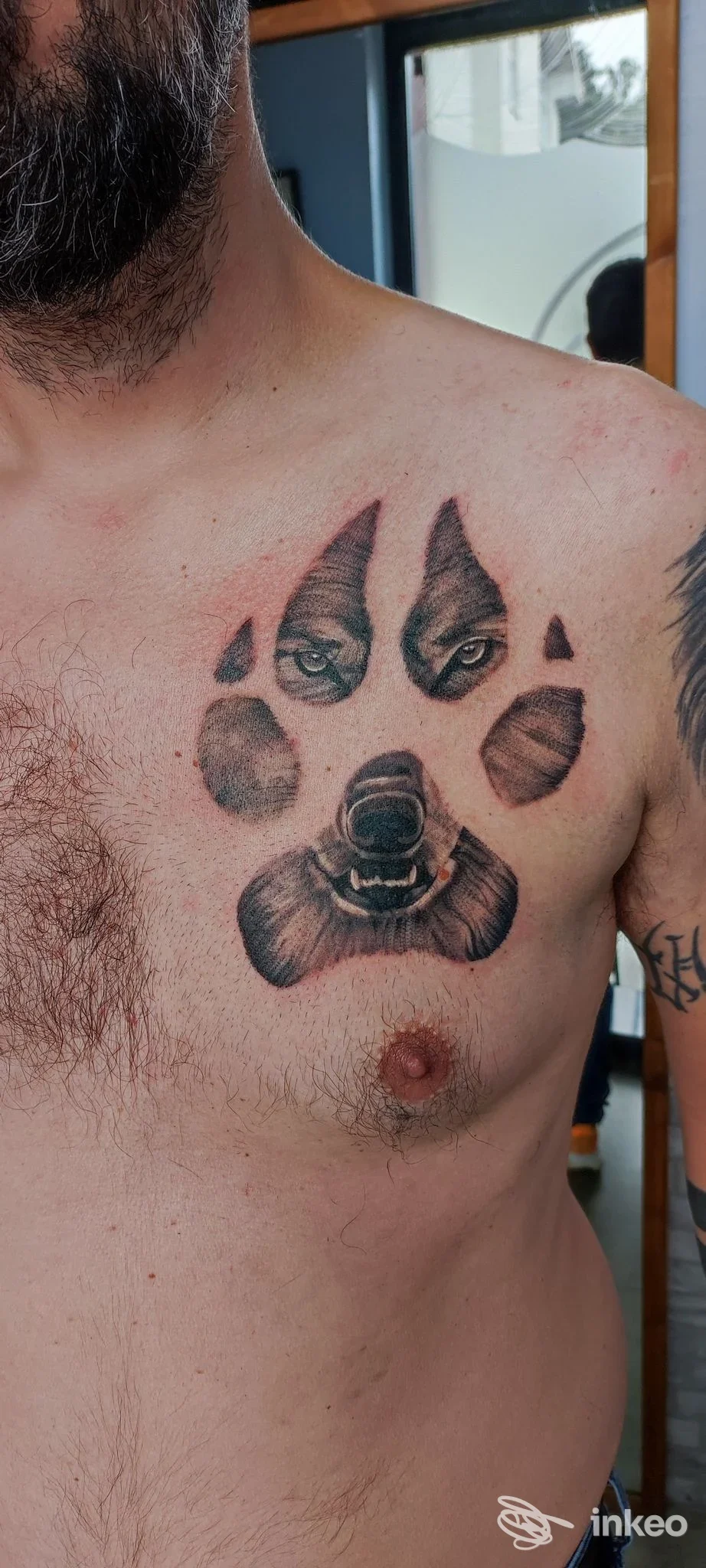 Tattoo sul torace sinistro raffigurante una zampa con una faccia di animale al centro: due occhi, naso e bocca, realizzato in tonalità nere e grigie.