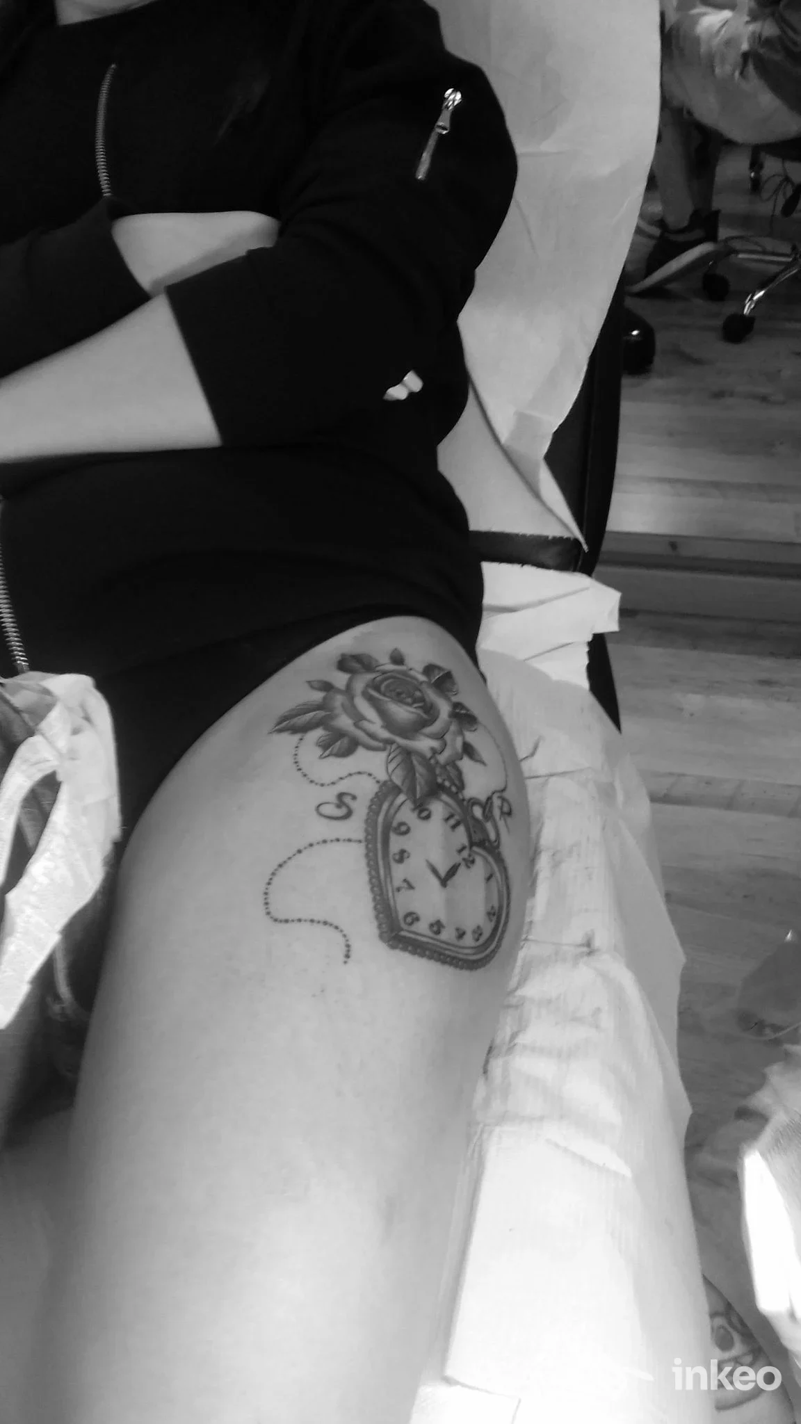 Tattoo realistico su coscia raffigurante un orologio a pendolo contornato da una rosa, visibile su una persona distesa in studio, in bianco e nero.