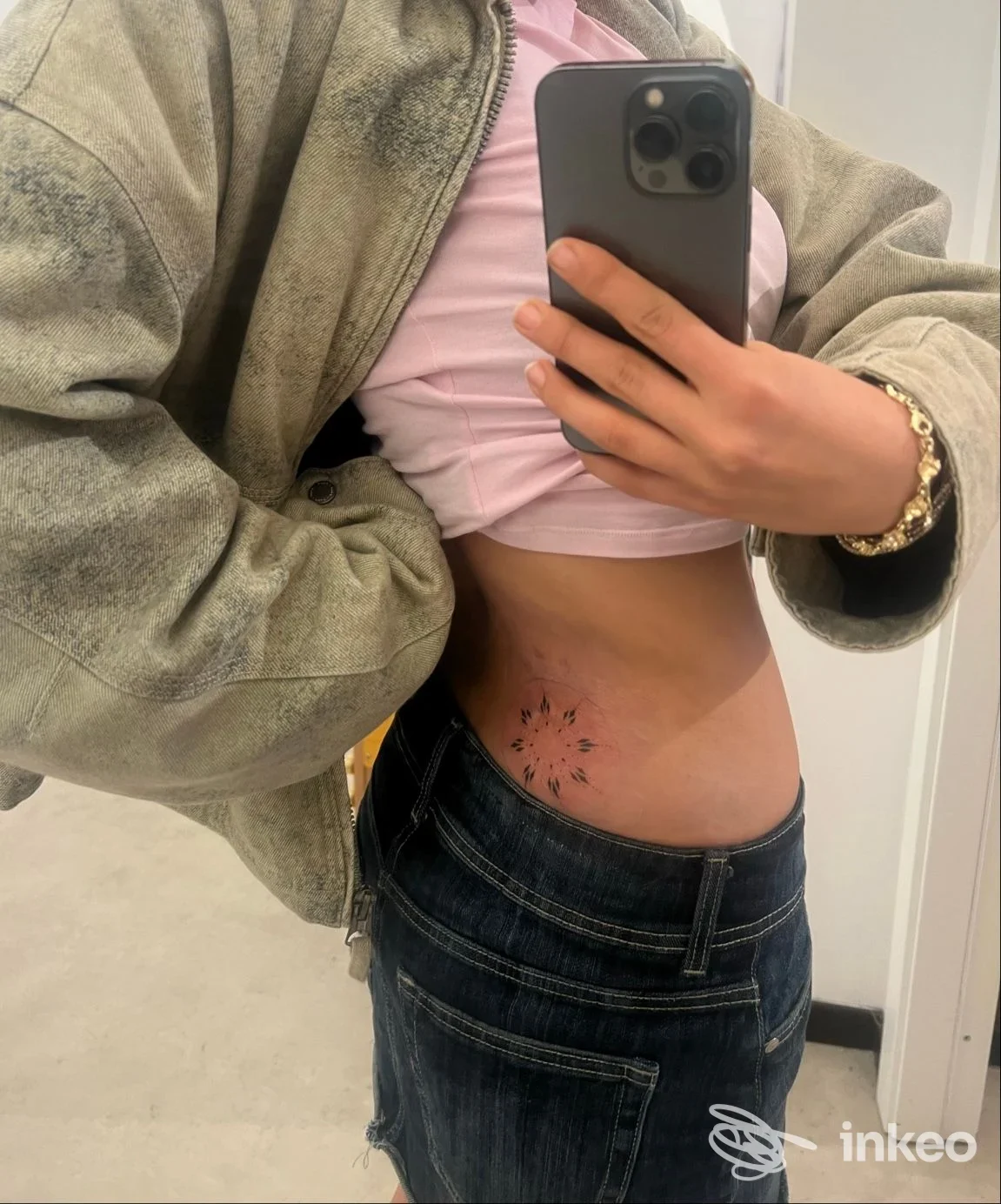 Soggetto con maglietta rosa sollevata che mostra un piccolo tatuaggio floreale lungo la costola sinistra, visibile sul fianco. Nessun volto visibile.