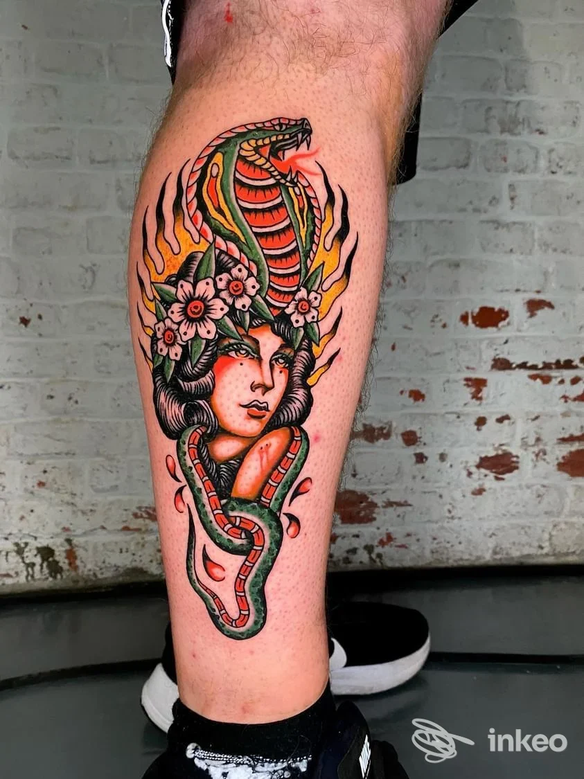 Tattoo colorato di una donna avvolta da un serpente con fiori, lungo il polpaccio, su pelle reale, parete in mattoni sullo sfondo.