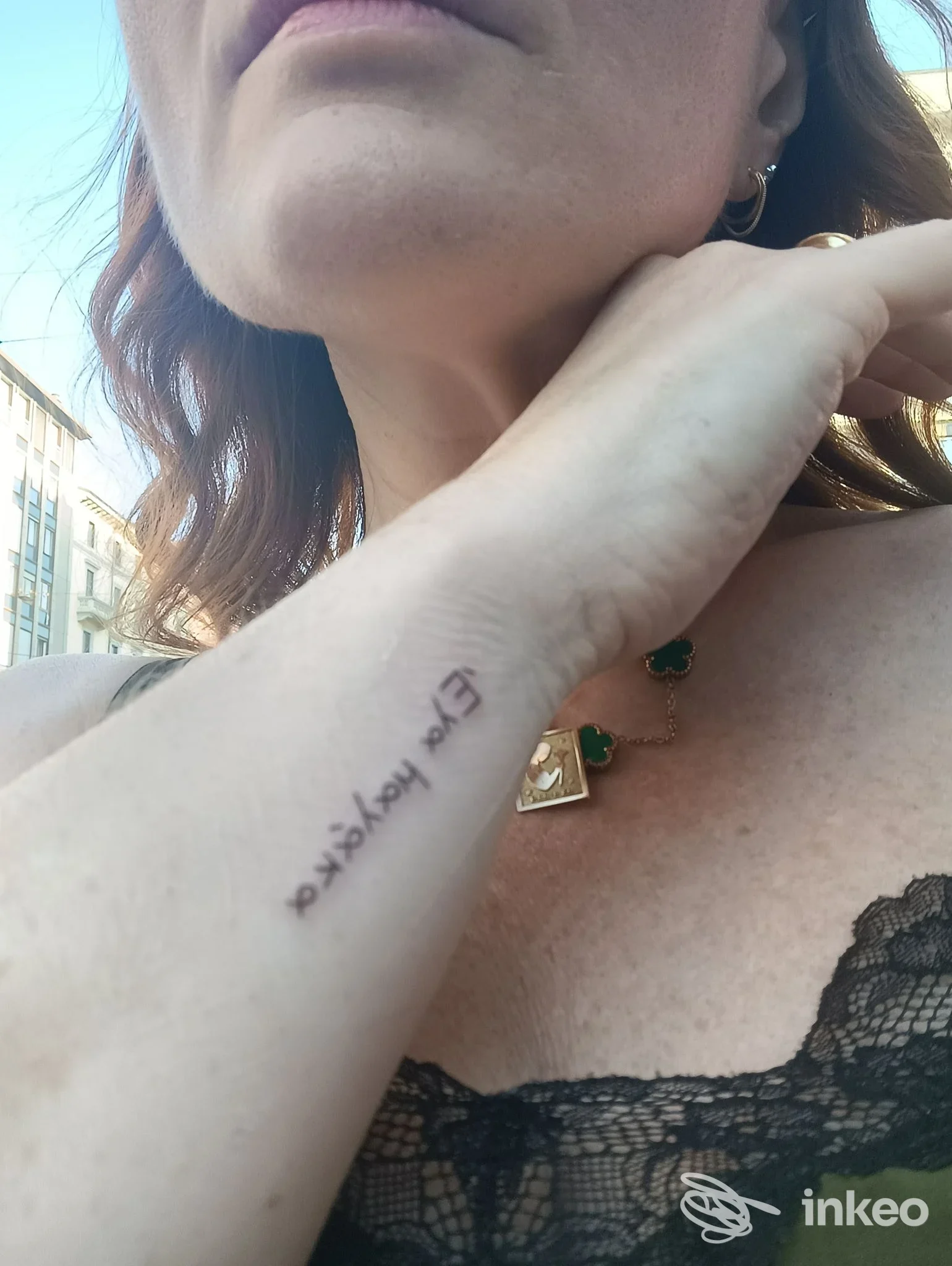 Primo piano di un braccio sinistro con tatuaggio testuale visibile sulla pelle, seguita da un collier verde e da un top in pizzo. Không watermark.