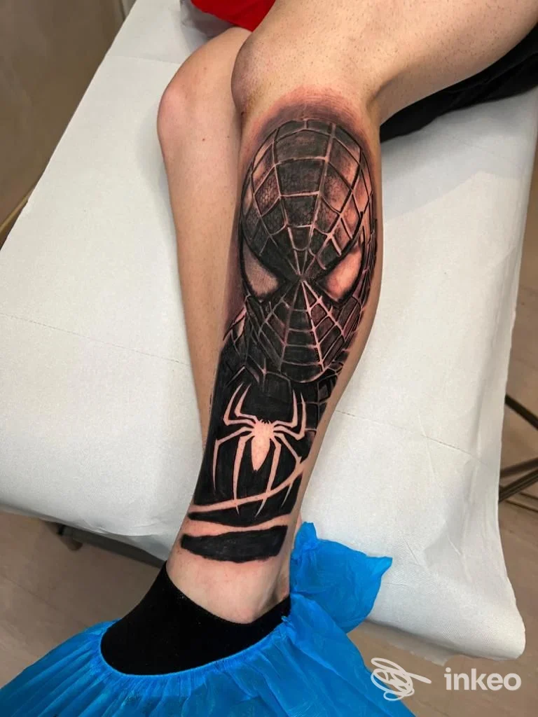 Tattoo sul polpaccio raffigurante Spider-Man, ritratto realistico su pelle, con tonalità nere e rosse, visibile una gamba di una persona seduta su lettino dal tatuatore.