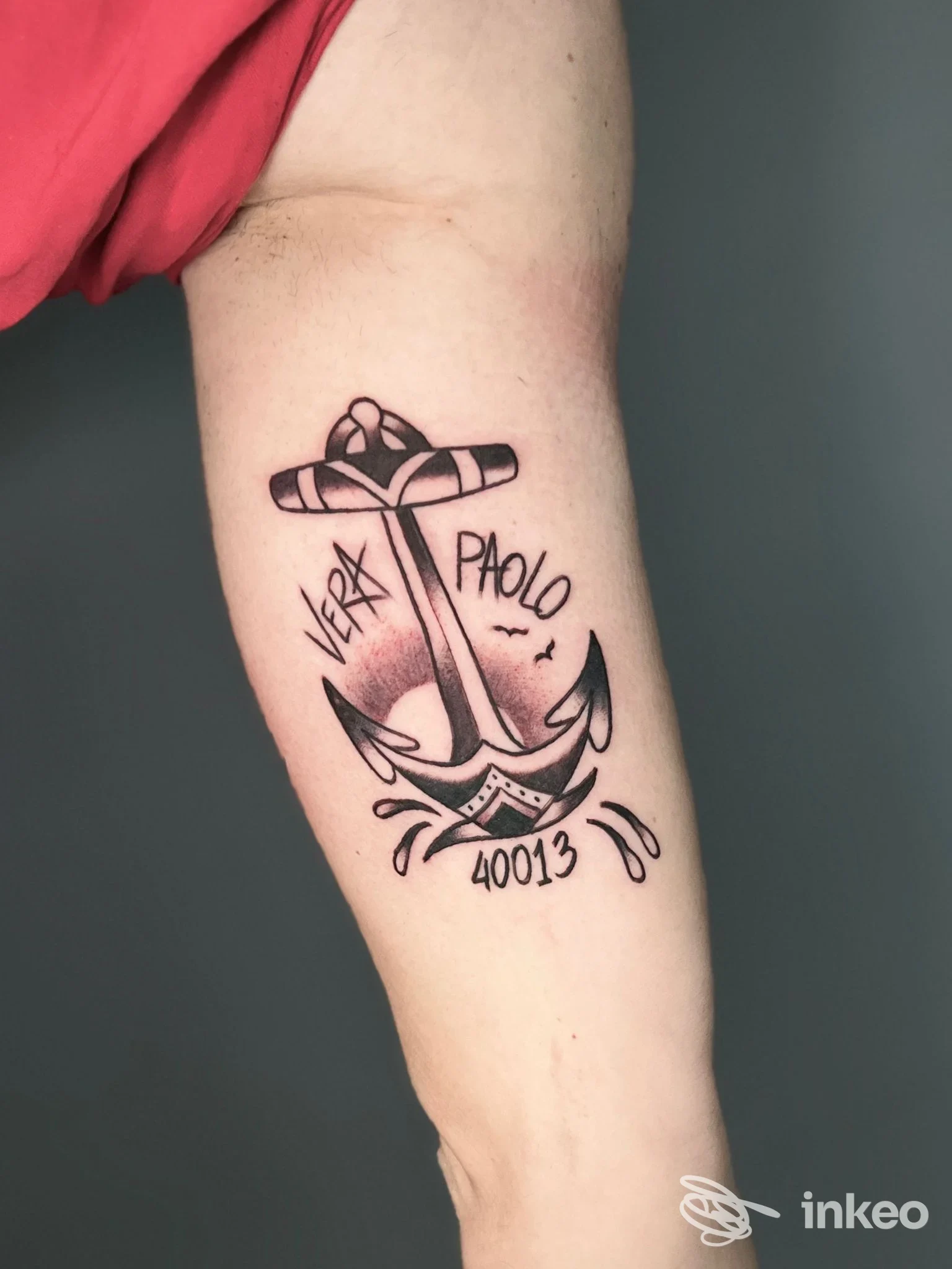 Tattoo di ancore e testo su braccio, linee scure e ombreggiature; visibile solo la parte di braccio, senza volto.