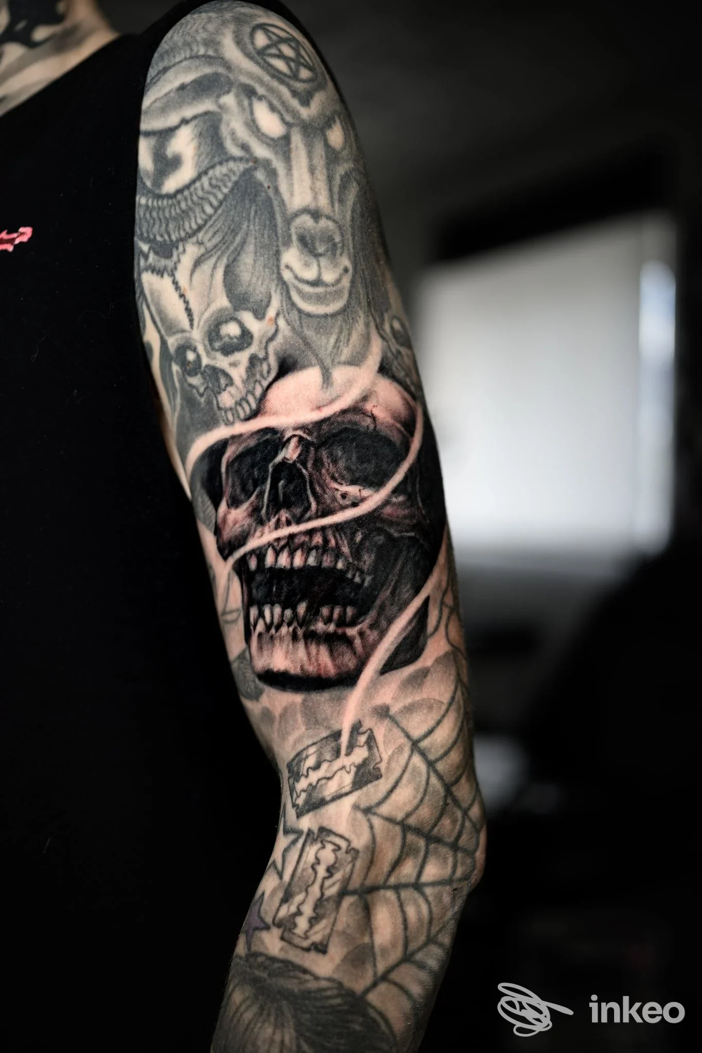 Braccio sinistro con un tattoo a manica scuro raffigurante teschi e motivi gotici, visibile in primo piano con sfondo sfocato.