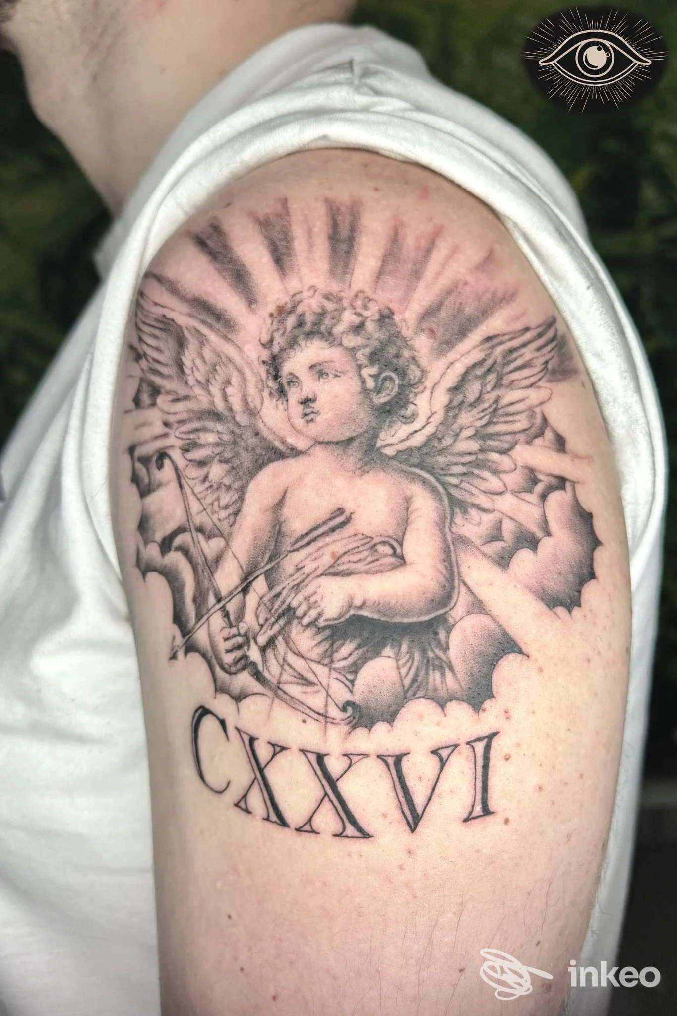 Tattoo realistico di un angelo cherubino con ali su spalla; numeri romani CXXVI sotto, realizzato sulla pelle di un braccio di una persona.
