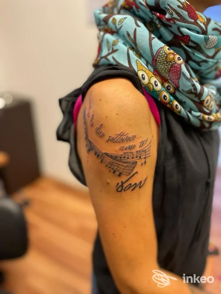 Tattoo sull'avambraccio visibile: linee e note musicali accompagnate da testo stilizzato, indumento colorato e sfondo neutro.