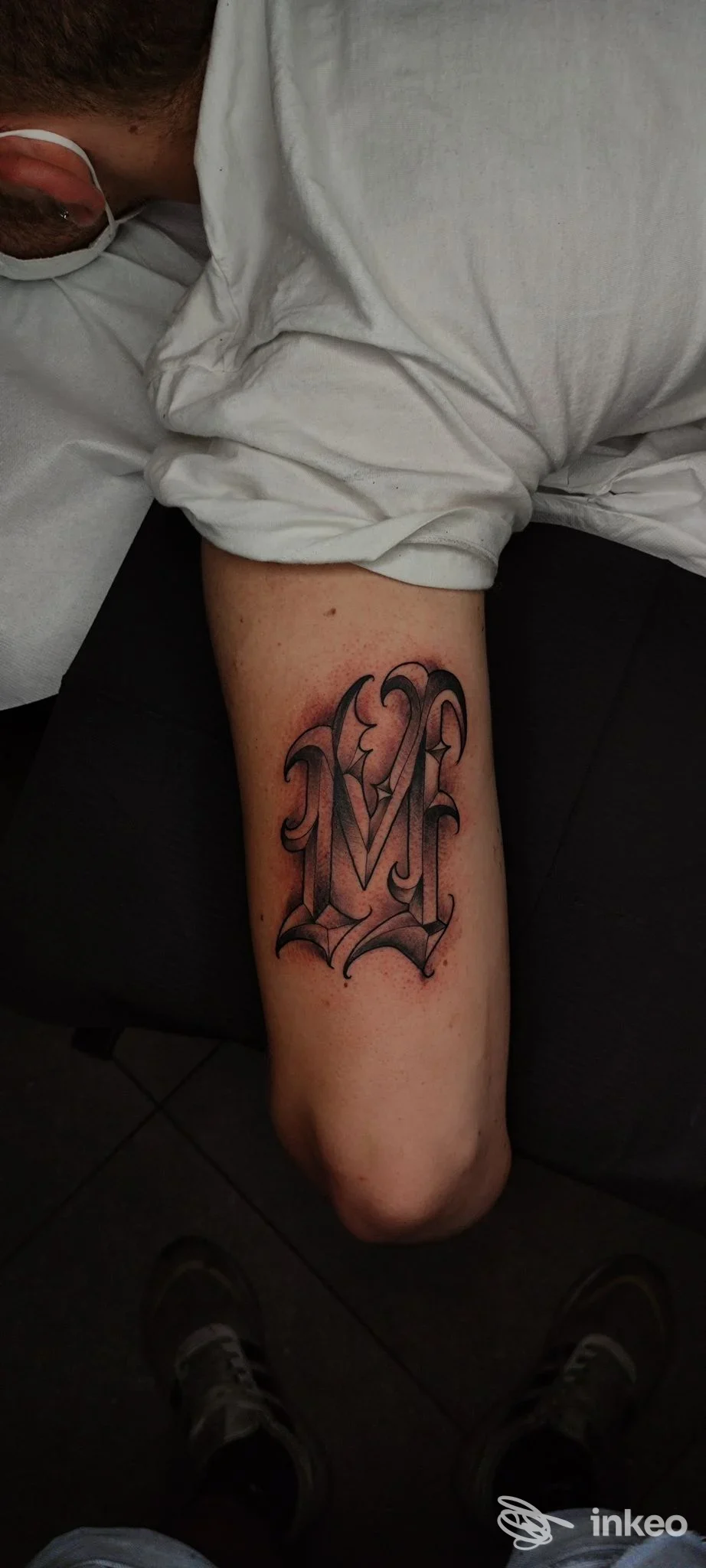 Tattoo visibile sull’avambraccio di una persona, stile ornato con lettere gotiche, colori scuri su pelle chiara, senza volto visibile.