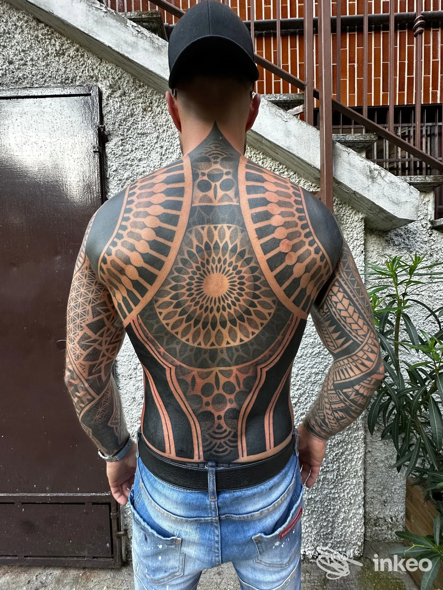 Foto di spalle di una persona con tatuaggio geometrico completo sulla schiena, visibile dalla nuca ai glutei, indossa cappellino e jeans.