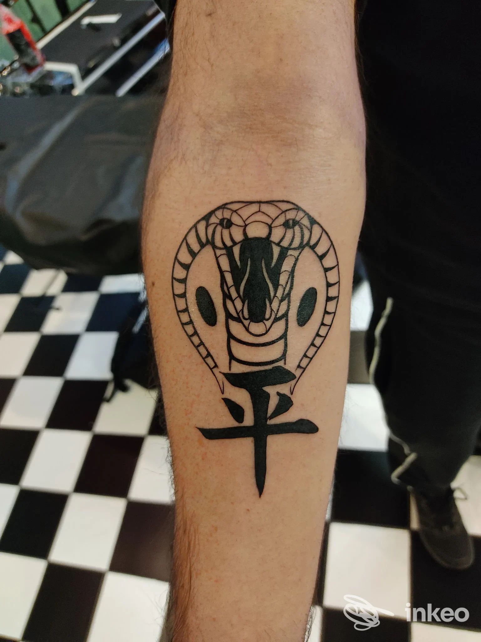 Avambraccio con tatuaggio: cobra stilizzato con cappuccio e simbolo sotto, realizzato sulla pelle umana, sfondo a pavimento a scacchi.