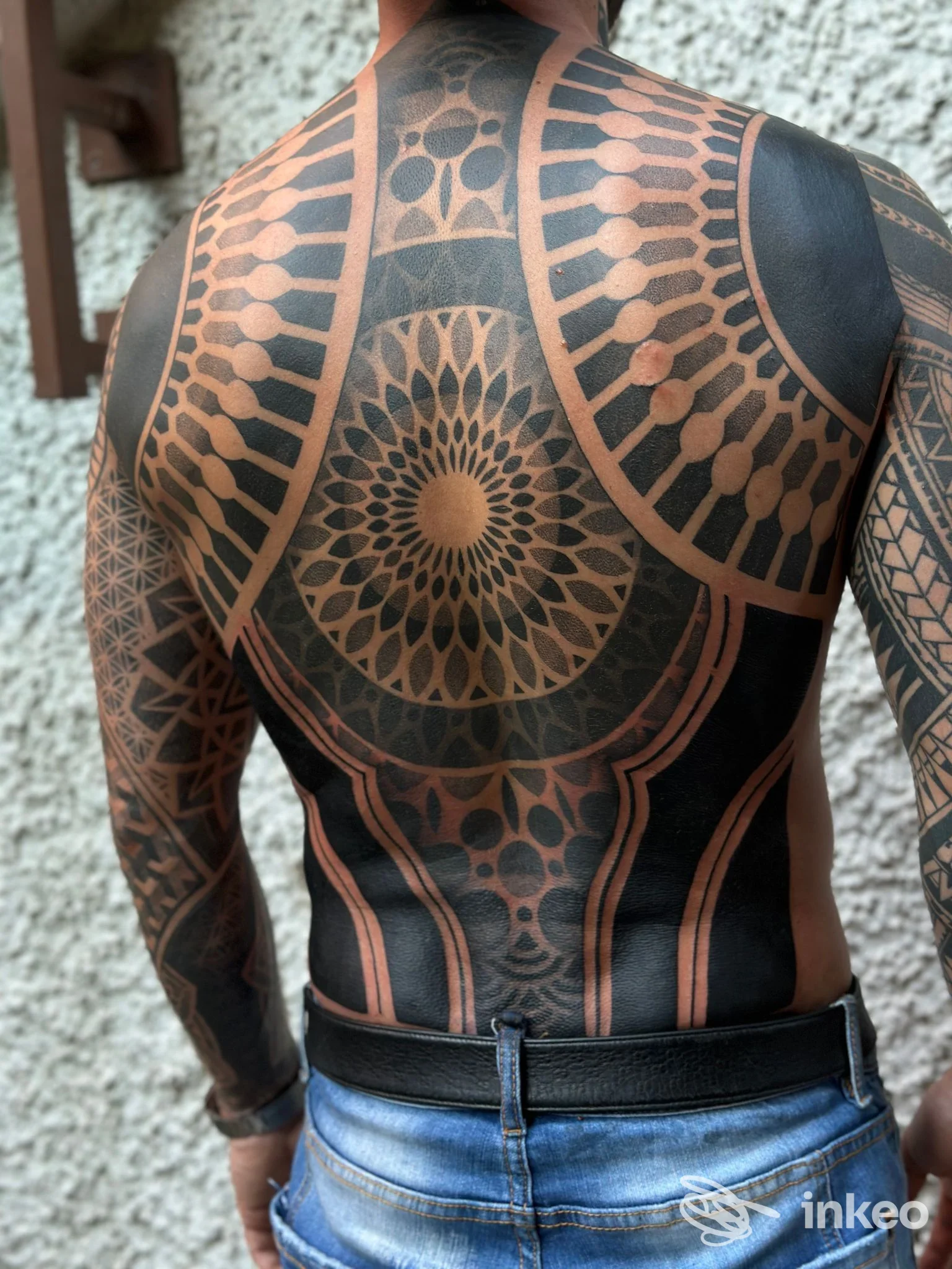 Sulla schiena di una persona si distingue un grande tattoo geometrico in nero con motivi circolari e linee che scorrono lungo la schiena; si intravedono le braccia coperte dall'opera.
