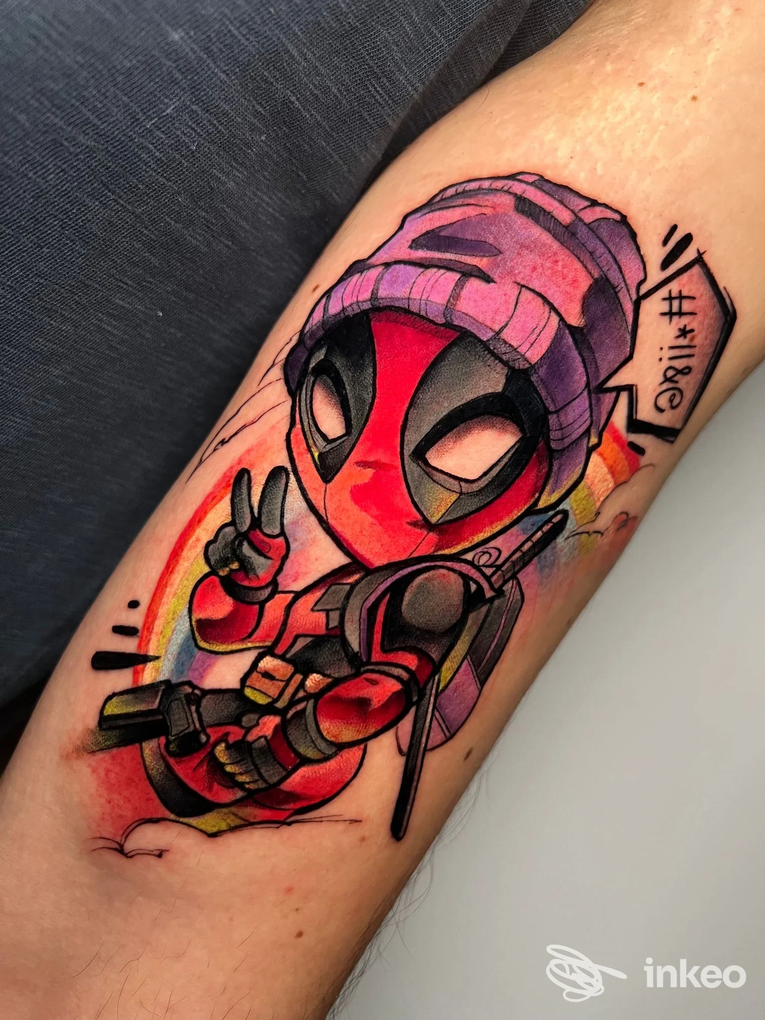 Tattoo sul braccio raffigurante Deadpool in stile cartoon, con berretto viola, realizzato in tinta vivace su pelle, in una singola inquadratura.