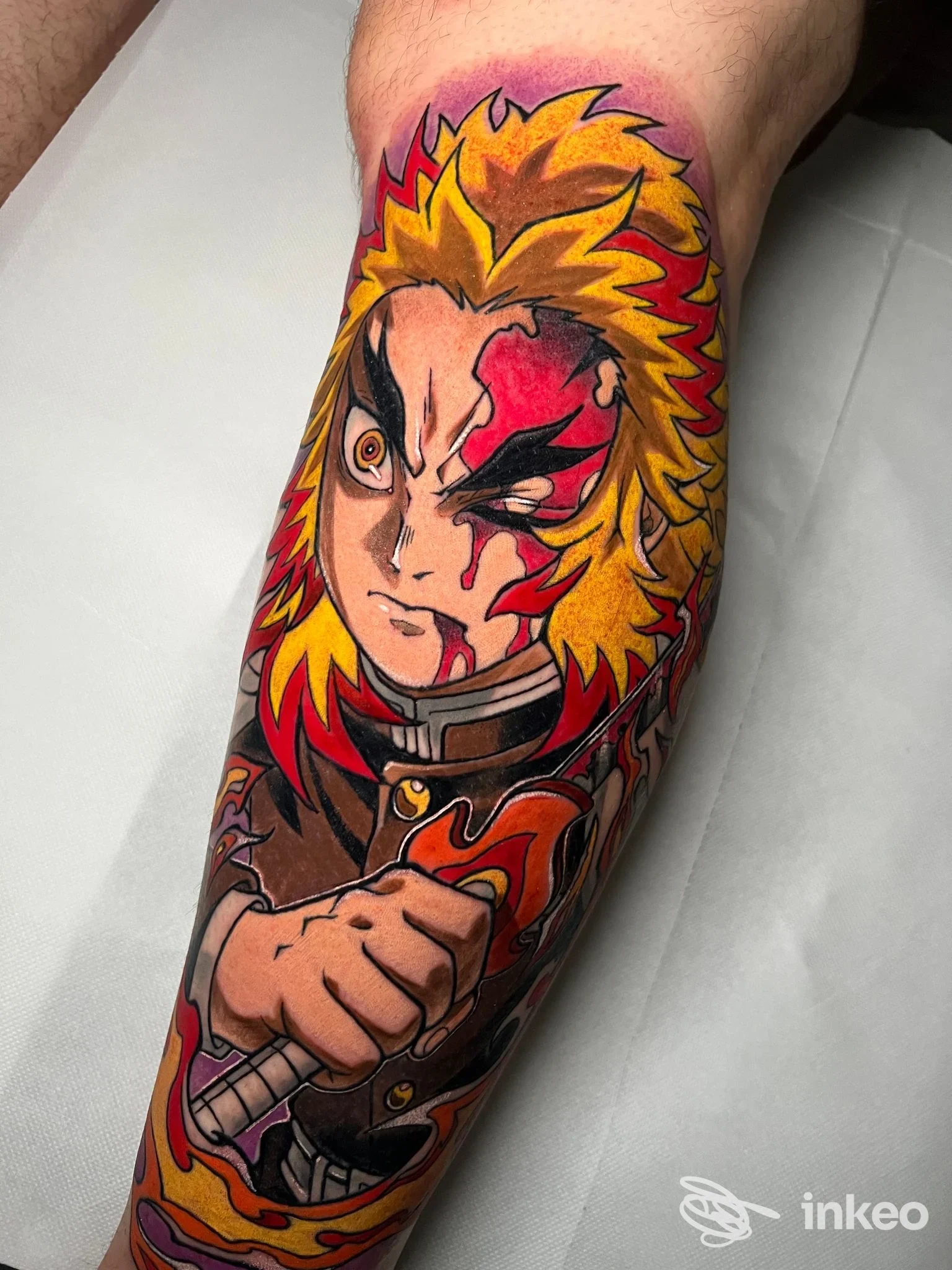 Tattoo a tema anime sul braccio sinistro, scena frontale del personaggio con capelli dorati e sfondo fiammeggiante, realizzato sulla pelle umana.