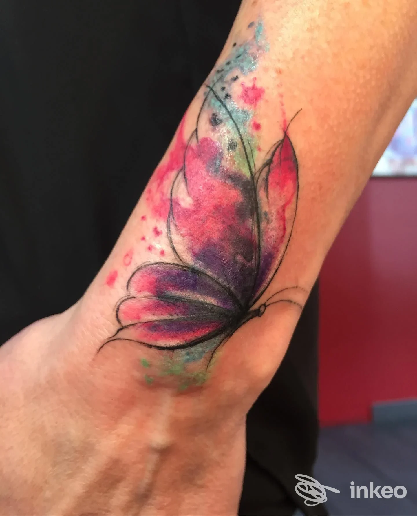 Tattoo sull'avambraccio raffigurante una farfalla in stile acquerello con sfumature rosa e viola, contorni neri sottili e schizzi di colore.
