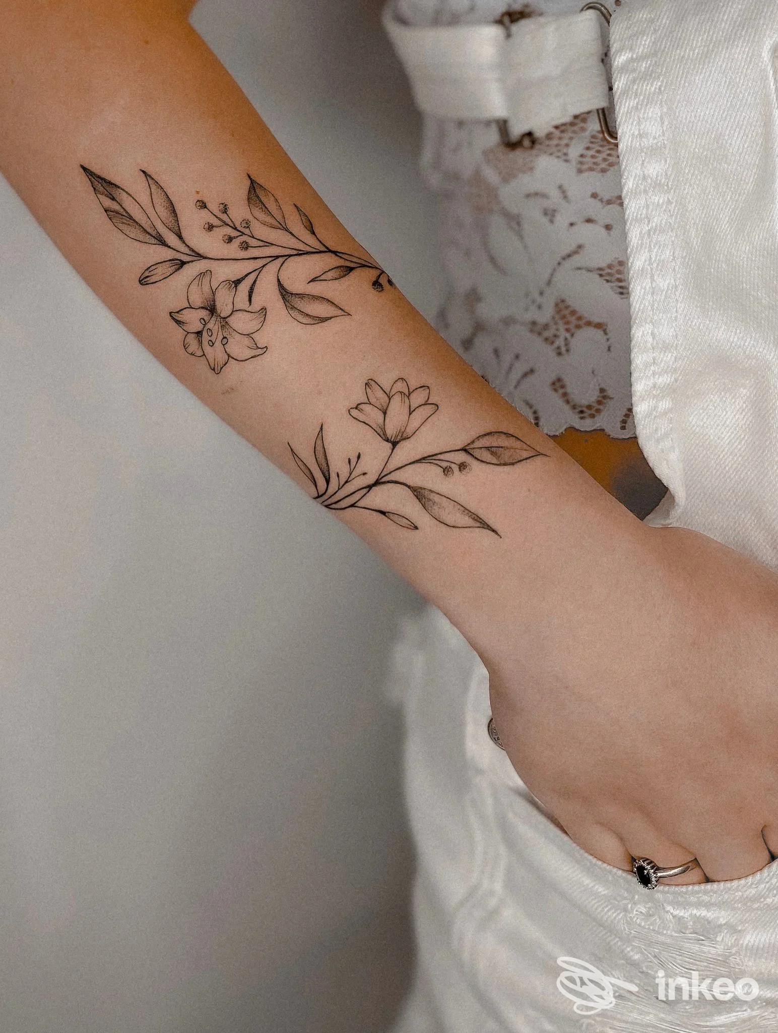 Tattoo floreale lungo l’avambraccio eseguito su pelle chiara, disegno a linea nera di fiori e rami, con una mano delicatamente appoggiata. Studio jey-tattooing-studio, città non indicata.