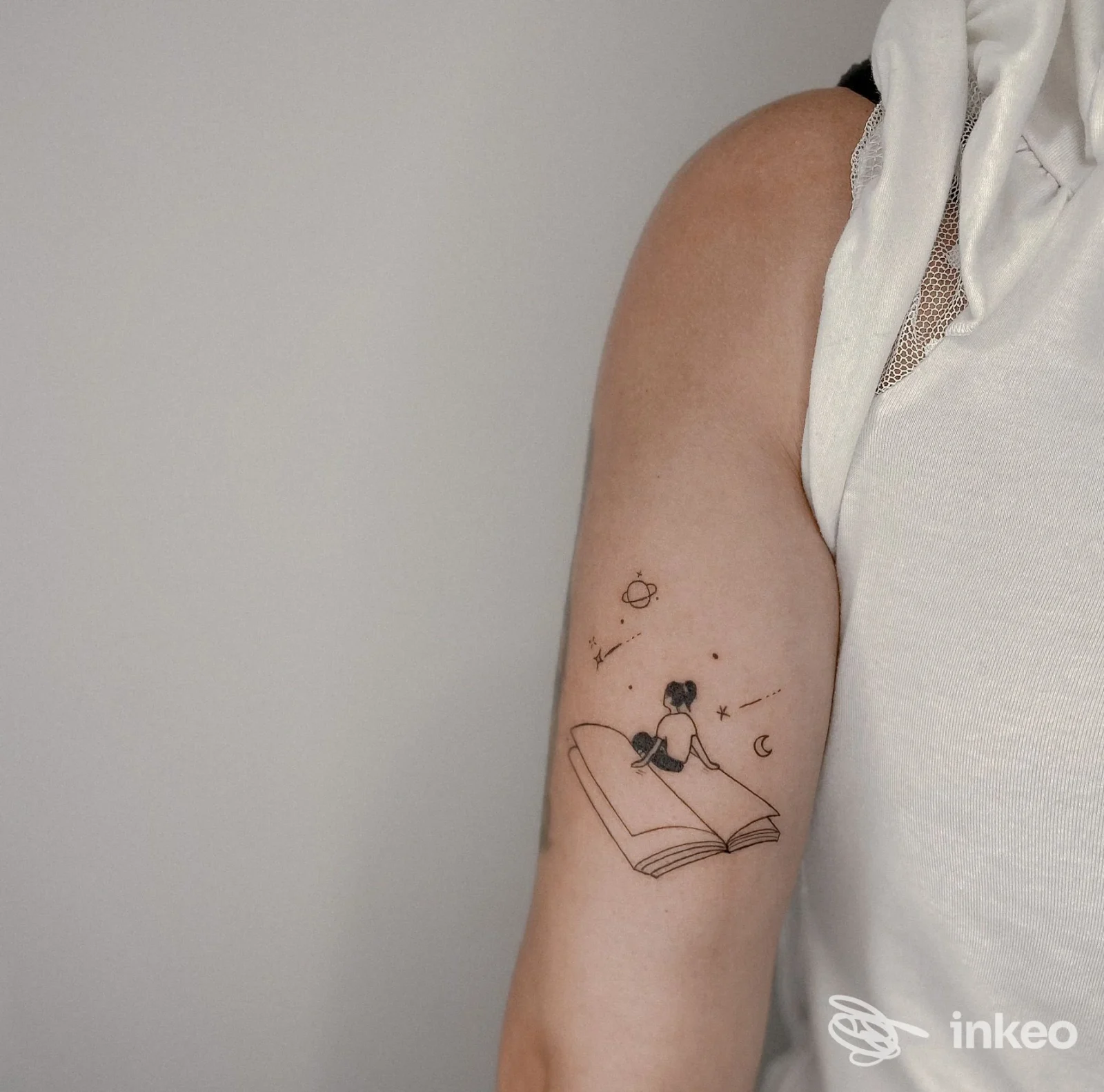 Tatuaggio visibile su avambraccio raffigurante una persona seduta su un libro aperto con elementi cosmici, sfondo neutro.
