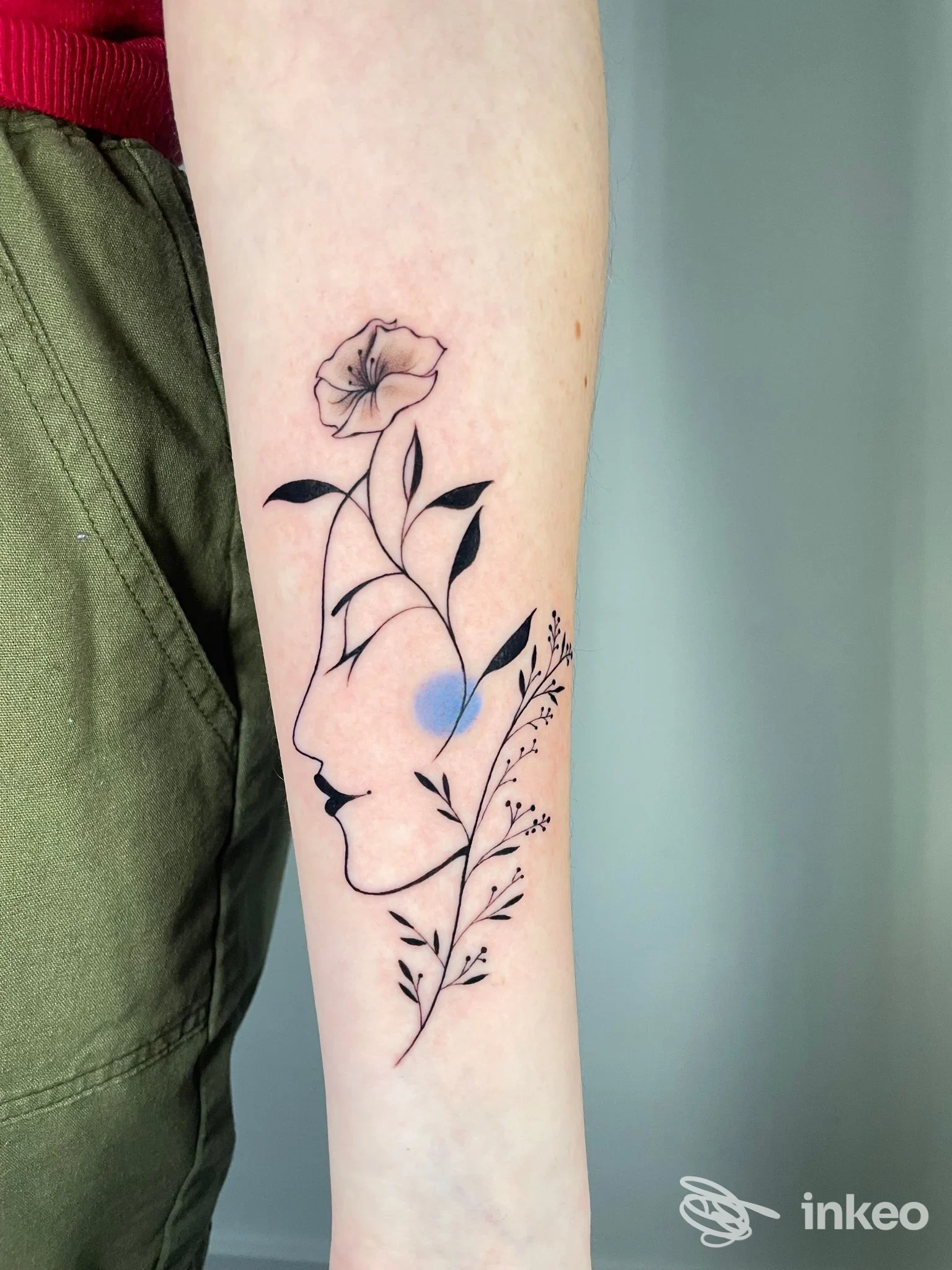 Tatuaggio floreale stilizzato sull'avambraccio: linea nera con rosa in alto, foglie scure e ramoscelli decorativi, con una sfumatura azzurra nello sfondo.