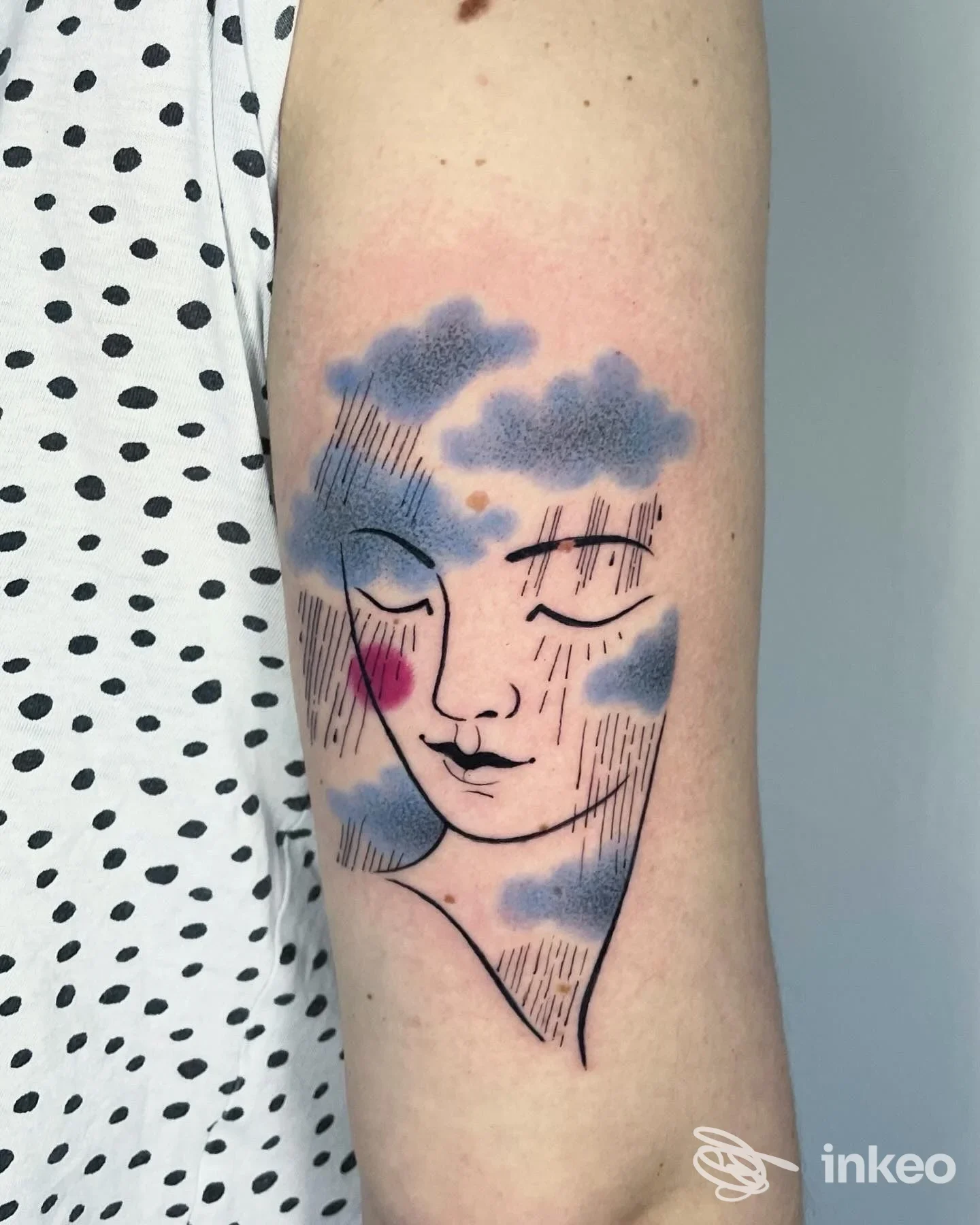 Tattoo su braccio raffigurante un volto stilizzato tra nuvole, con linee sottili e sfumature blu e rosa; l’immagine mostra solo la parte corporea interessata.
