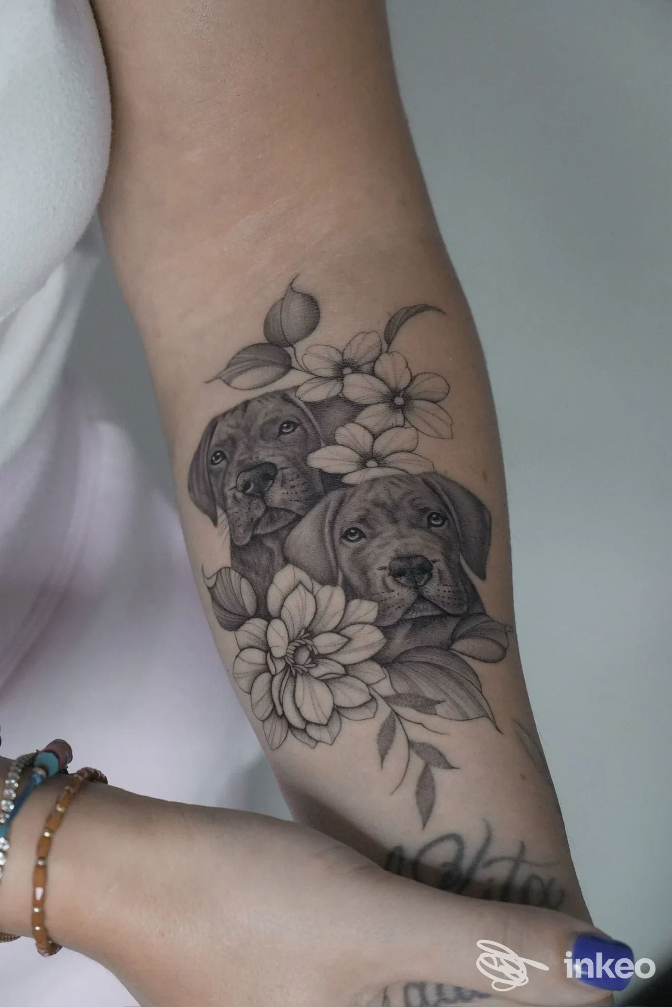 Tattoo inchiostro nero e grigio sull'avambraccio sinistro raffigurante due cani circondati da fiori, lavoro corporeo visibile e realizzato sulla pelle.