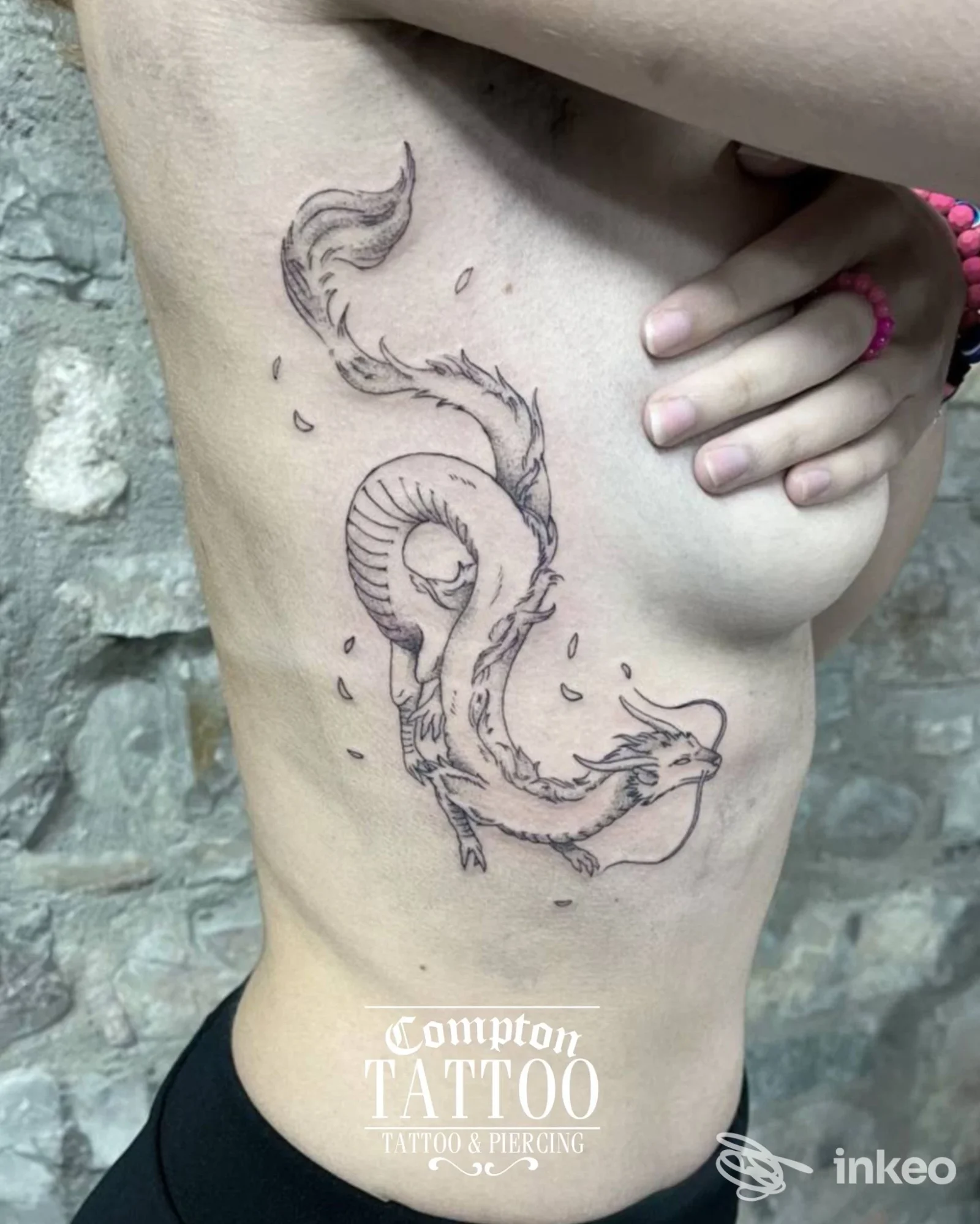 Tattoo su fianco sinistro di una creatura serpente-dragone, lineare e dettagliata, con parete di pietra sullo sfondo; parte del corpo umano visibile senza volto intero.