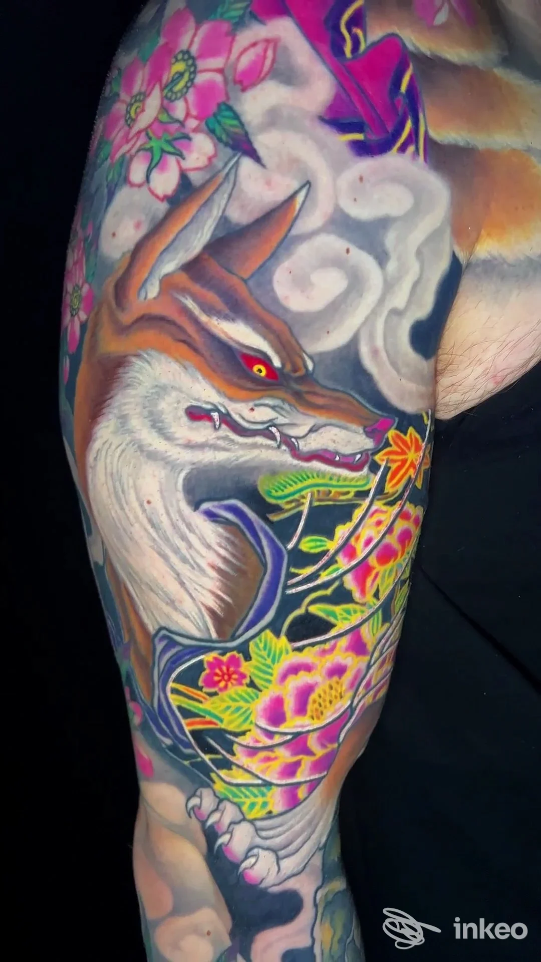 Tattoo colorato su braccio sinistro, creatura stilizzata con fiori rosa e gialli, sfondo scuro; senza volto umano visibile.