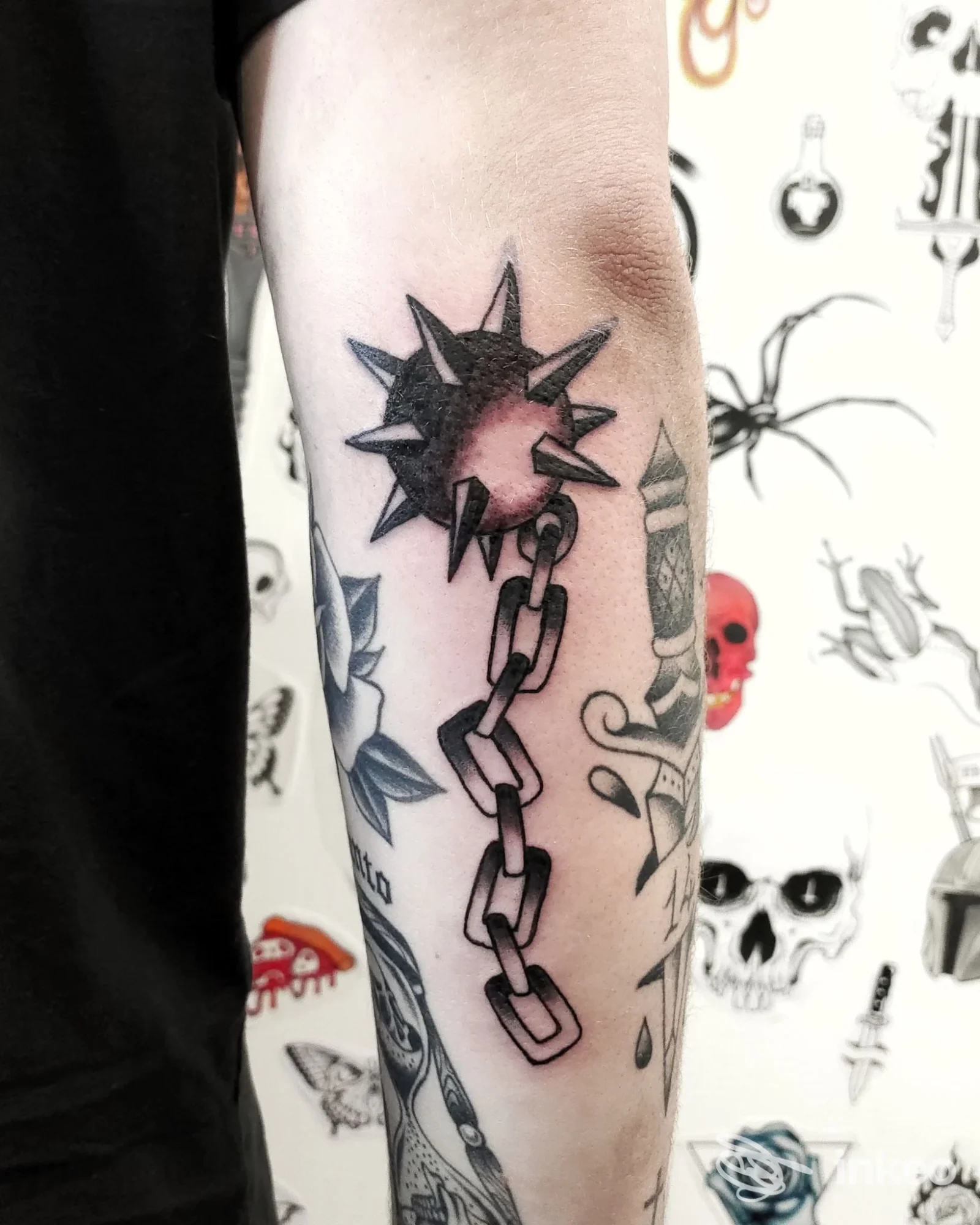 Tatuaggio sul braccio: stella spessa con punte irte collegata a una catena, realizzato sulla pelle dell’avambraccio, con altri disegni visibili sullo sfondo.