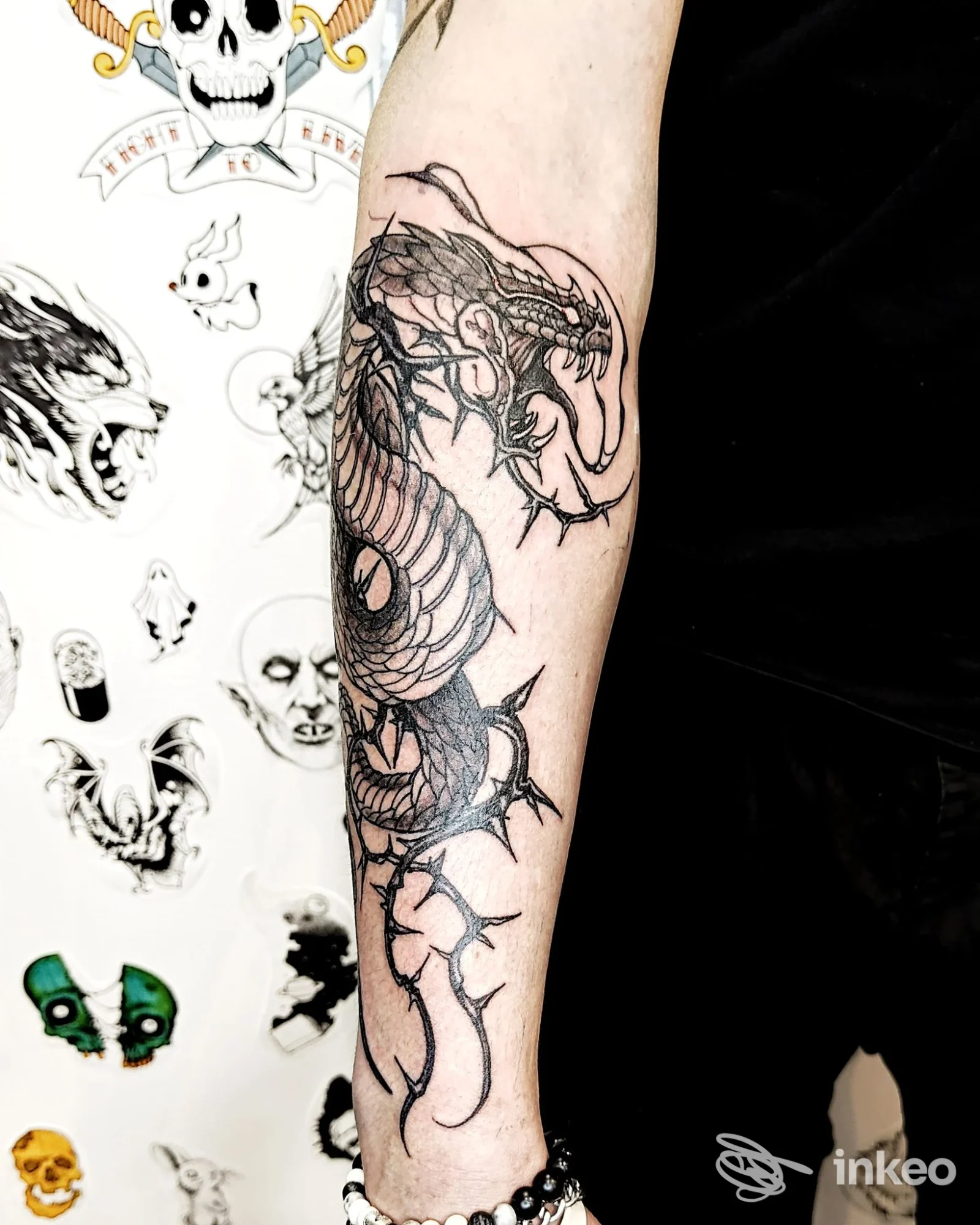 Tattoo drago su avambraccio sinistro, disegno a linea nera su pelle chiara, sfondo con disegni su muro.