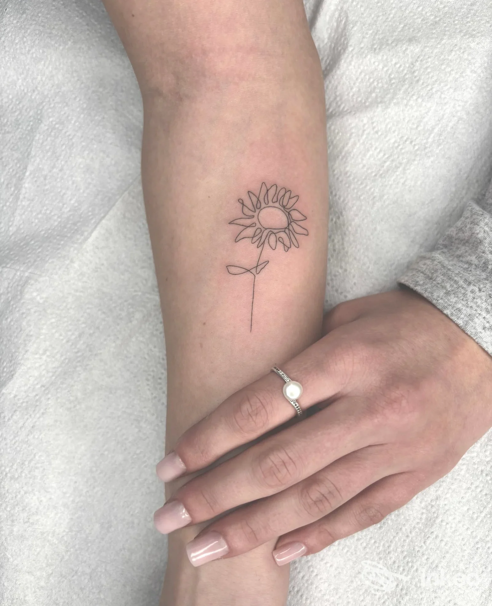 Tatuaggio minimalista a linea raffigurante una girasola sul braccio, eseguito direttamente sulla pelle; la mano dell'utente è visibile accanto al braccio.