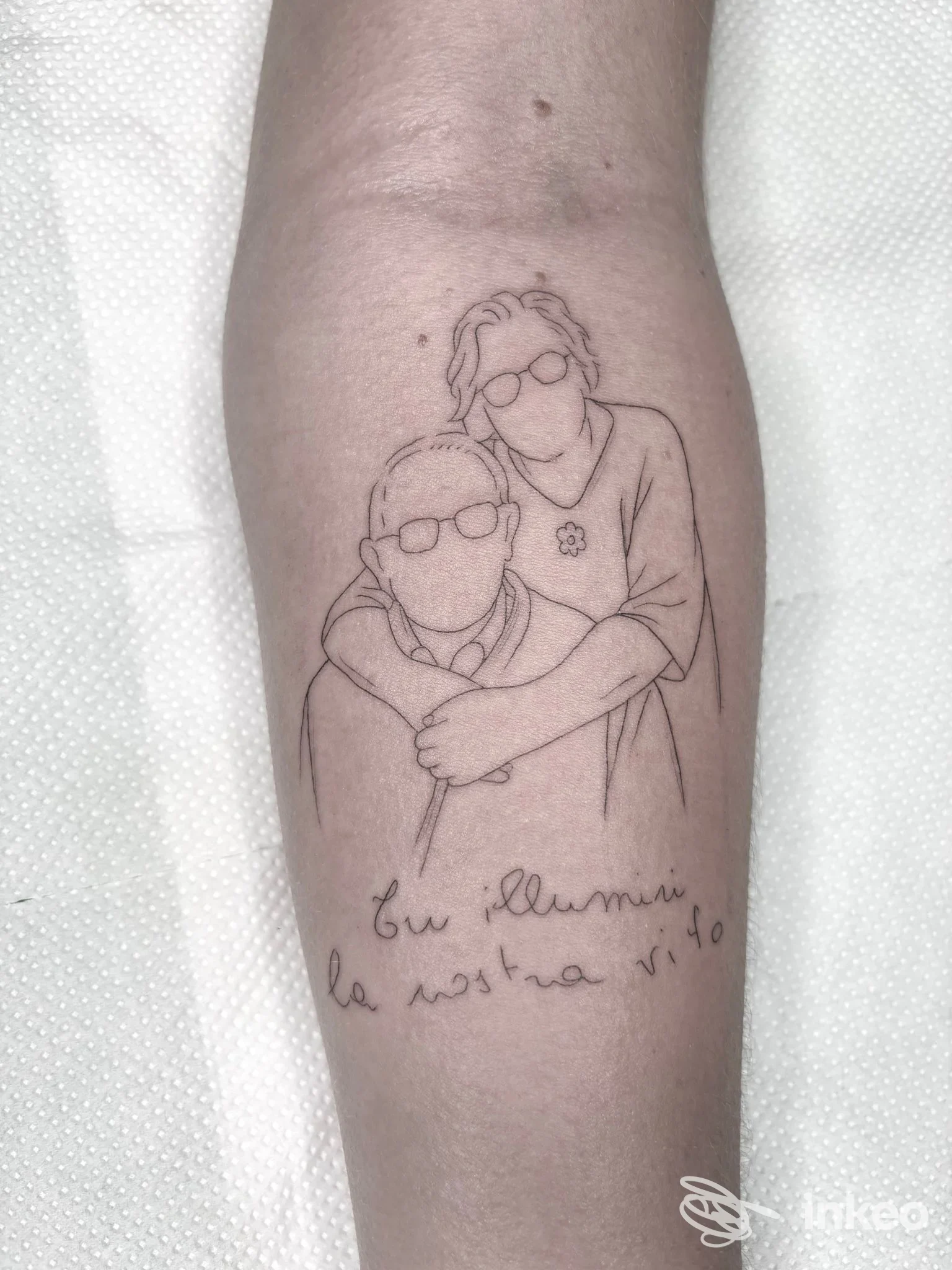 Disegno di tatuaggio lineare su avambraccio raffigurante due persone che si abbracciano, con testo in corsivo sul tatuaggio.