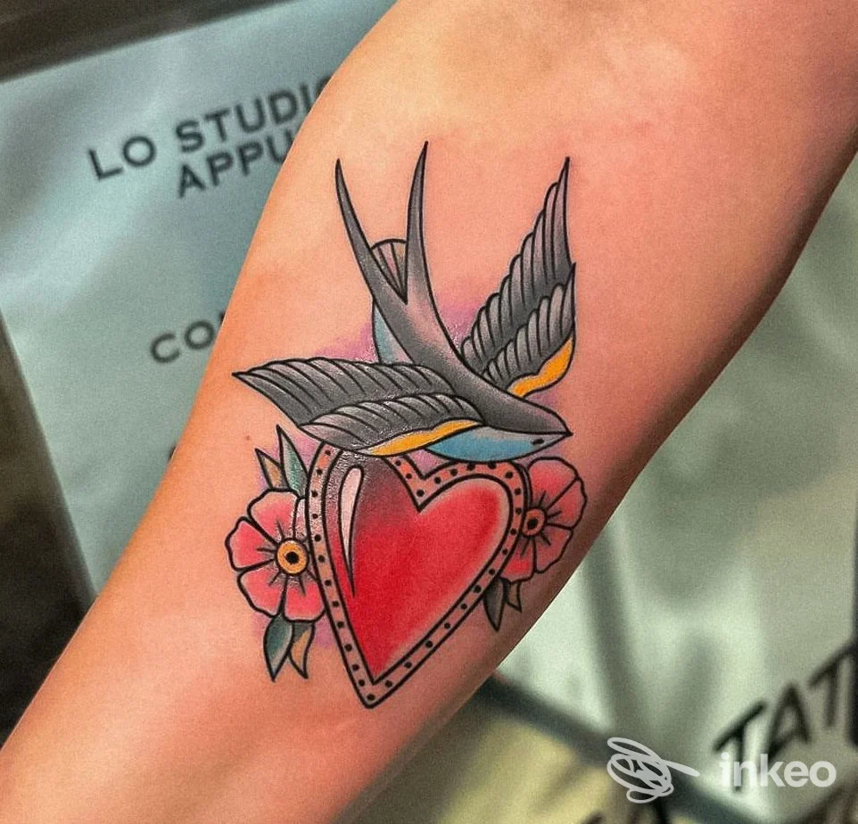 Tattoo sul braccio raffigurante un cuore rosso con rondine stilizzata e fiori, contorni netti e colori vividi.