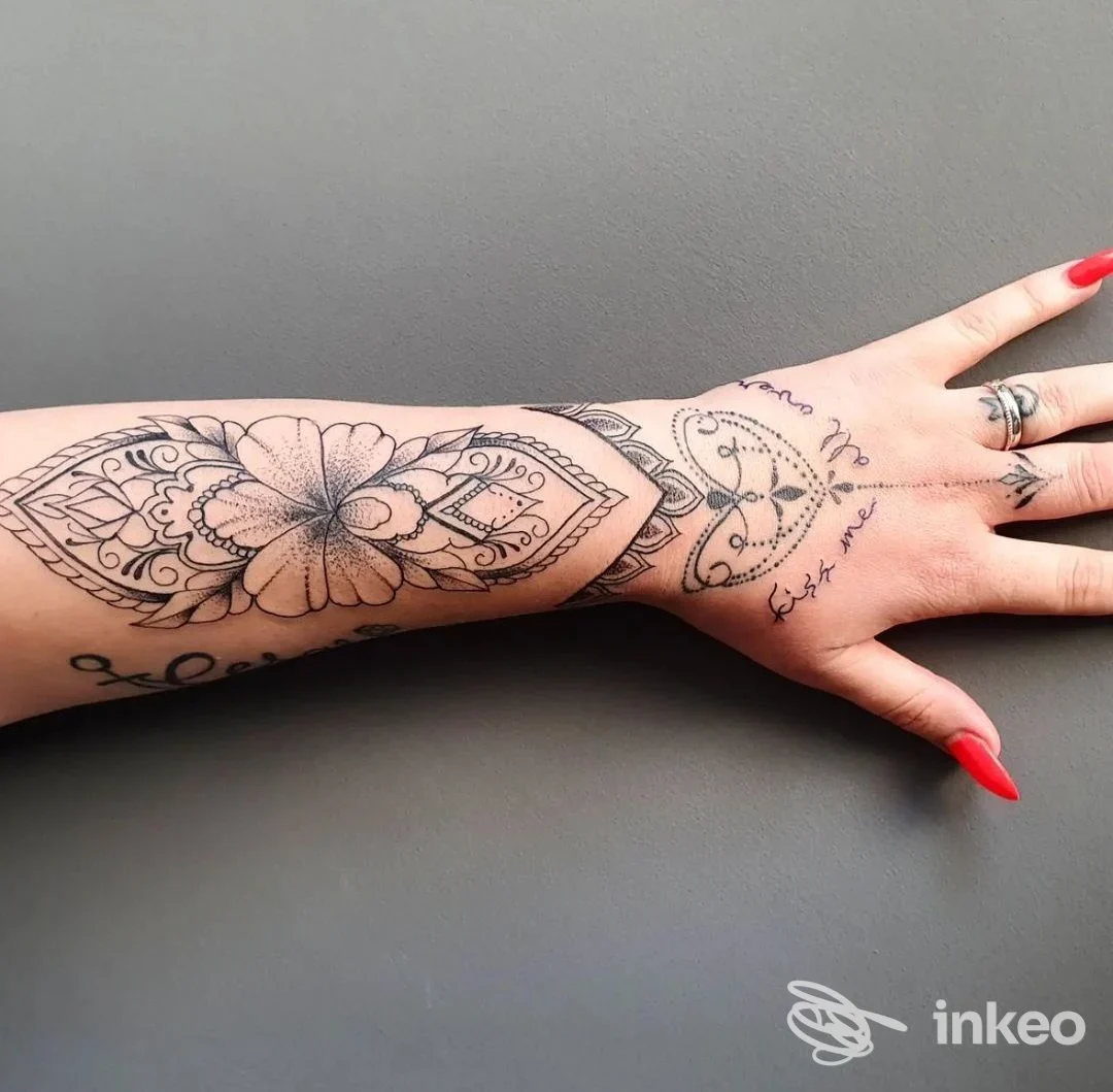Avambraccio e mano sinistra con tatuaggio geometrico e floreale, inchiostro nero su pelle chiara, con unghie rosse, sfondo grigio