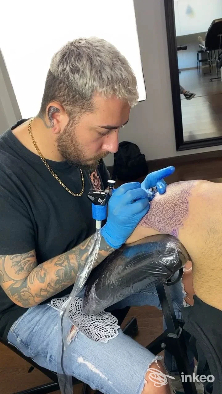 Un tatuatore lavora su una pelle, applicando una macchina per tatuare; guanti blu, posa concentrata, presso Stray Dog Tattoo Shop a Pescara.