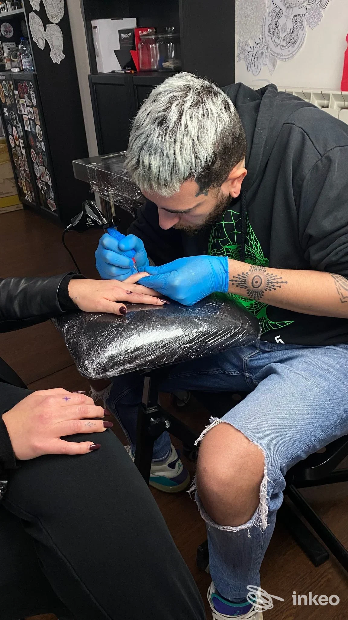 Un tatuatore con guanti blu lavora su una mano di un cliente in un negozio di tatuaggi, con sfondo di studio e pareti decorate a destra.