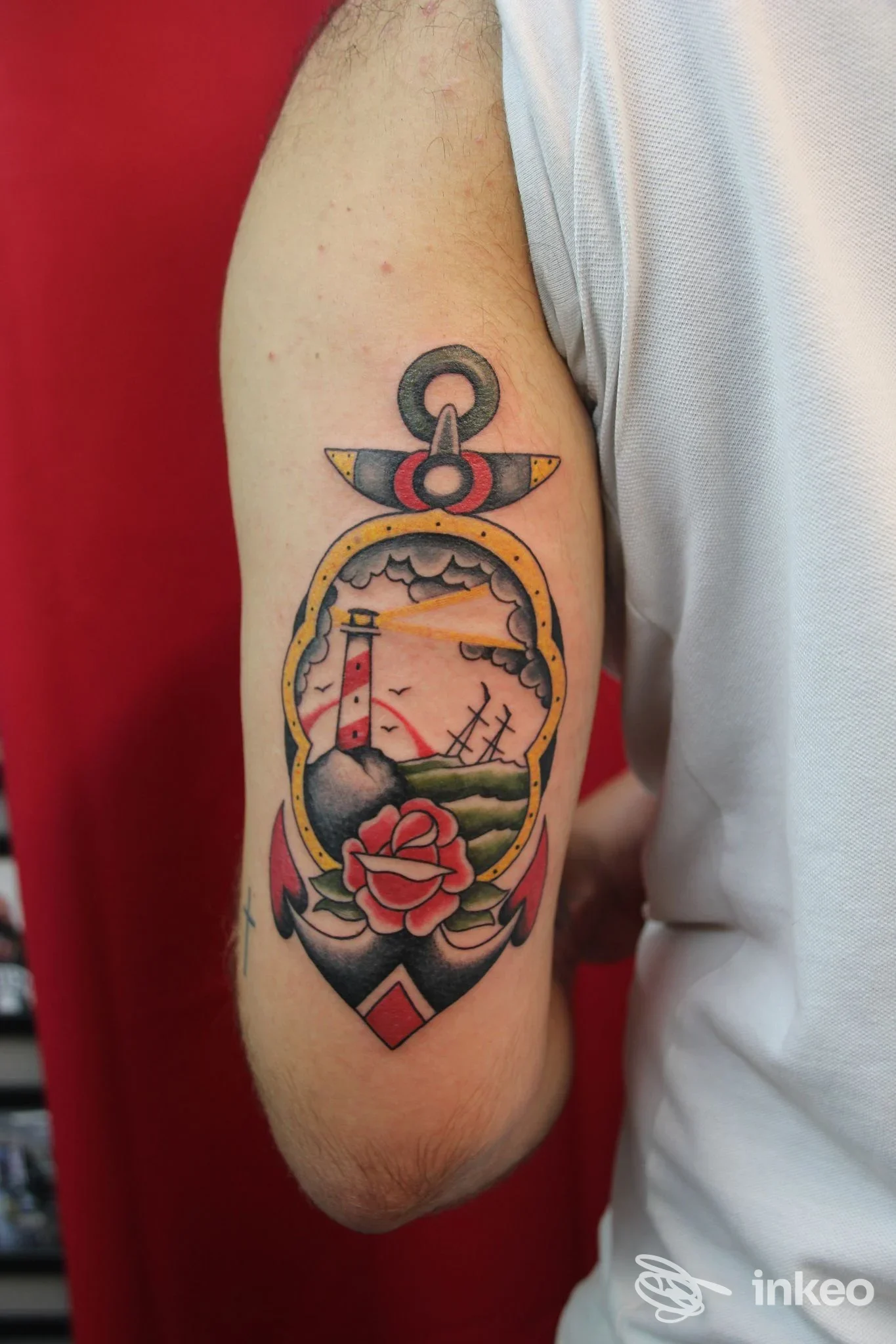 Tattoo sull'avambraccio sinistro: paesaggio con faro, mare e rosa, contorni neri e sfumature colorate, con un anello nautico in alto e decorazioni sul bordo.