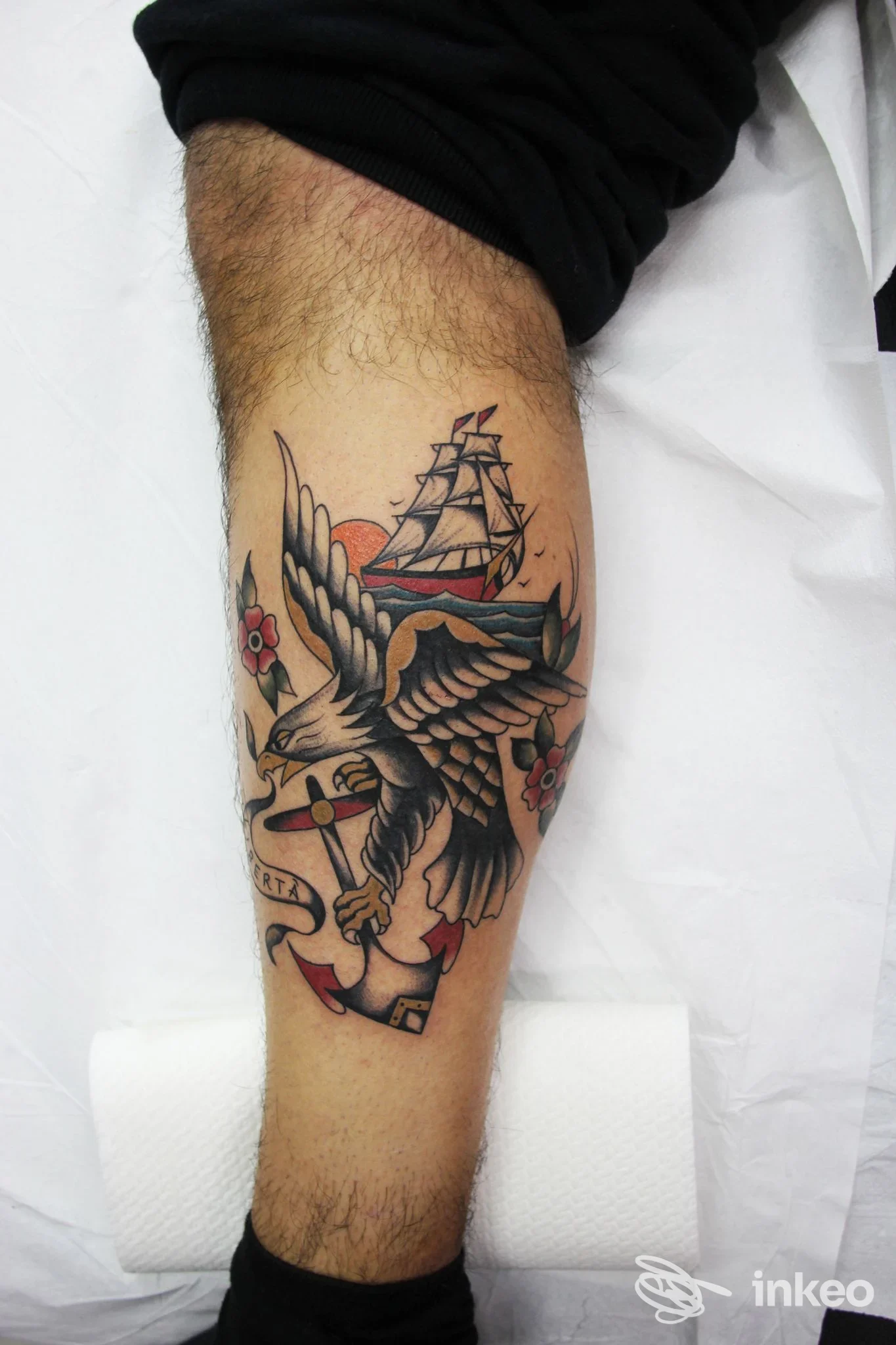 Tattoo su polpaccio raffigurante un uccello in volo con una nave e un'ancora in stile tradizionale, colori nero, rosso e marrone, visibile chiaramente sul tronco inferiore della gamba.
