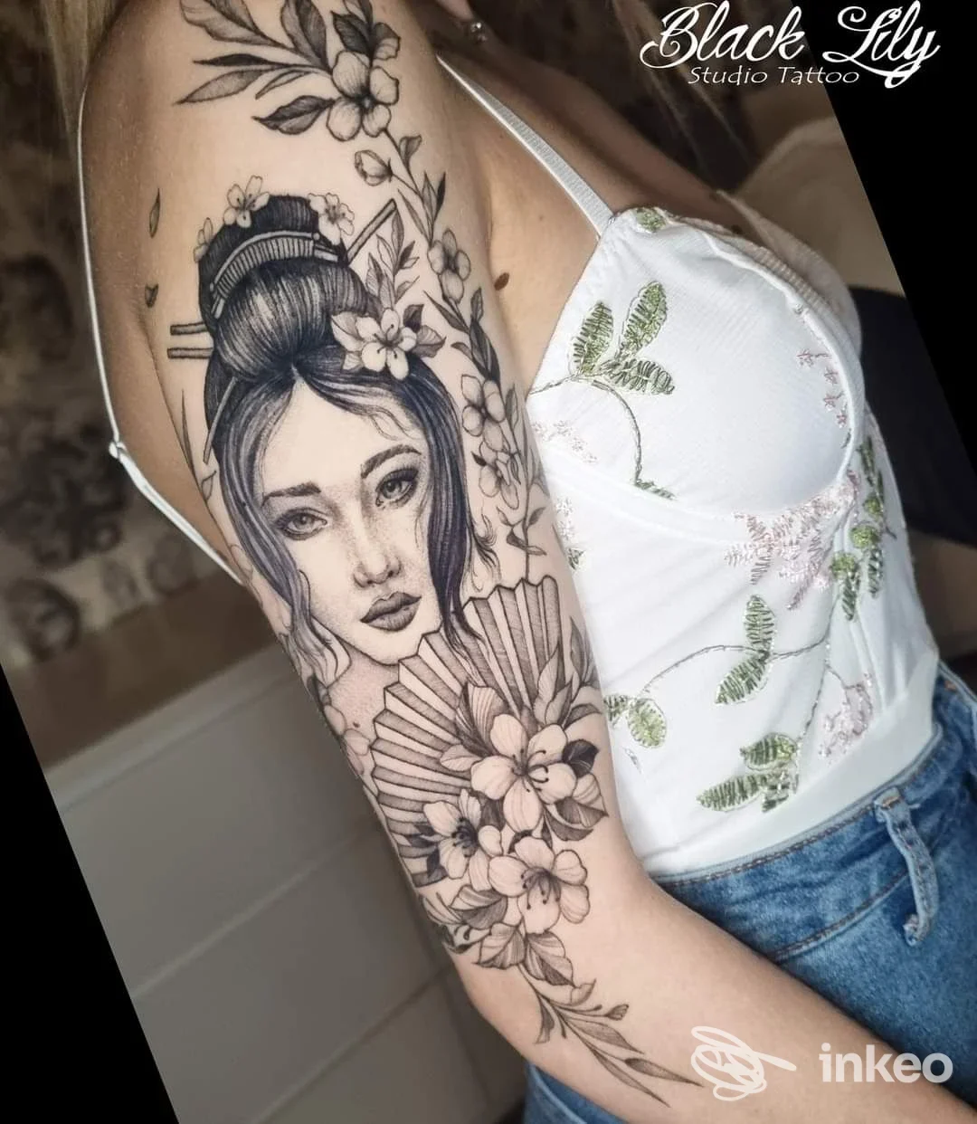 Tattoo su braccio raffigurante una geisha con corone e fiori, in stile black e grigi, realizzato su pelle della modella, scatto frontale parziale.