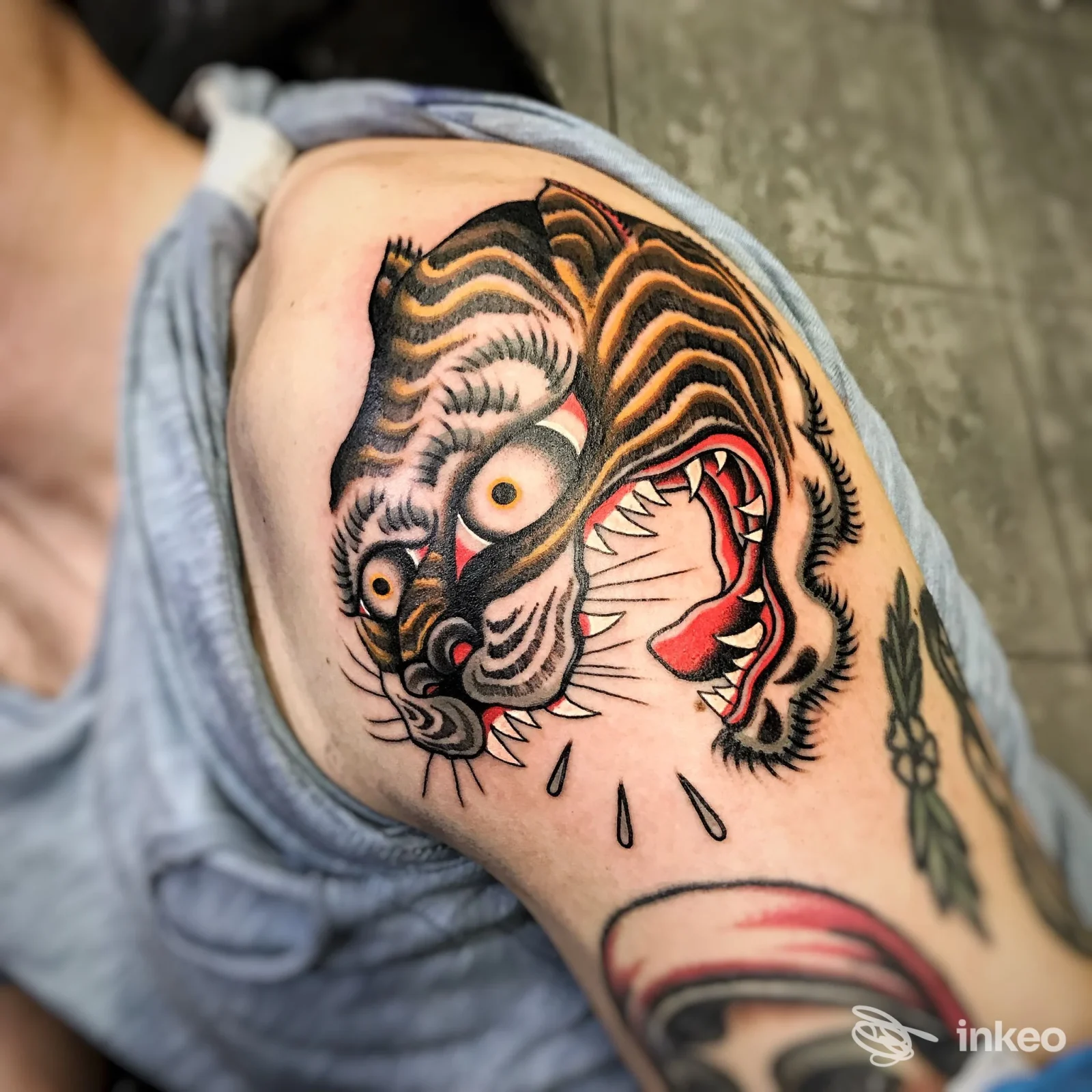 Tatuaggio rappresentante una tigre stilizzata sulla spalla destra, con linee marcate nere, sfumature arancioni e una bocca rossa molto aperta, visibile sulla pelle.
