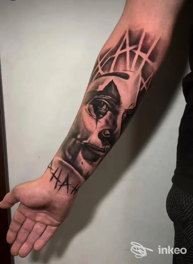 Avambraccio sinistro con tattoo realistico di volto e elementi geometrici sovrapposti; tonalità nero-grigie, senza overlay o testo visibile.