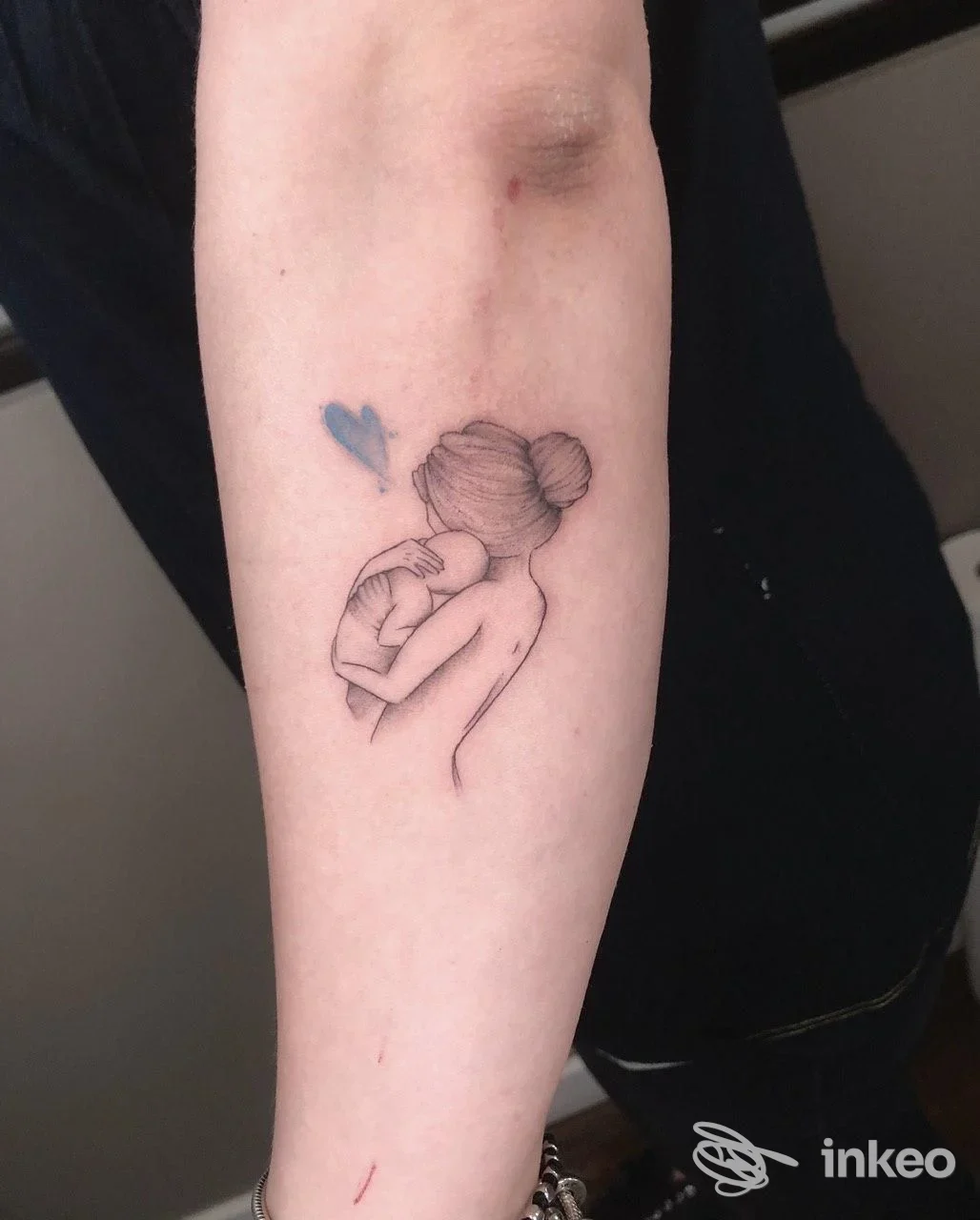 Tattoo sull'avambraccio: figura stilizzata di una donna che tiene in braccio un neonato, con un piccolo cuore blu accanto, imprecisamente visibile contro la pelle.