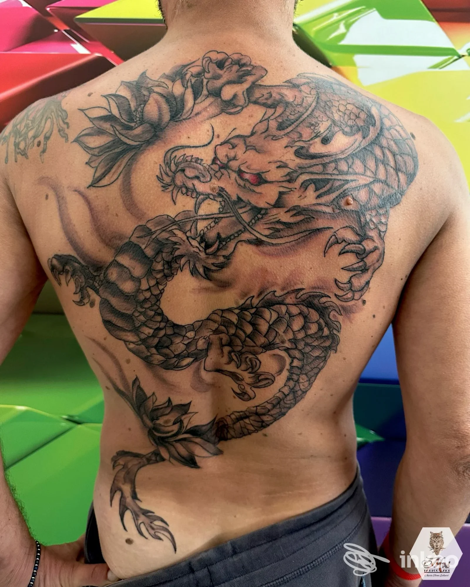 Schiena di un uomo con un grande tatuaggio drago in stile tradizionale, nero/grigio, visibile sulla pelle durante una sessione in studio.