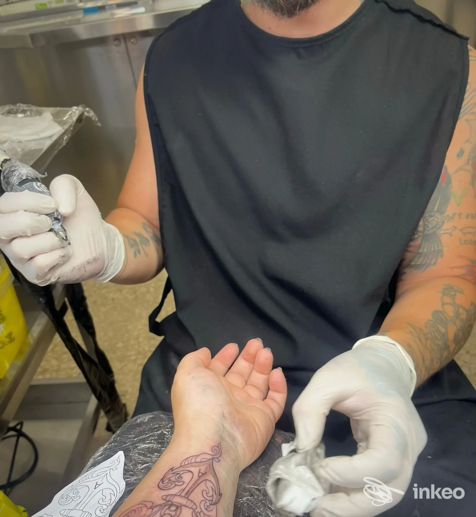 Primo piano di una sessione di tatuaggio: una mano con guanti tiene il telaio della macchina mentre un tatuatore lavora sull’avambraccio, con tatuaggio già visibile sulla pelle.