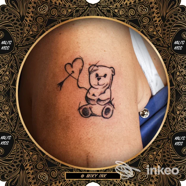 Tatuaggio su braccio raffigurante orsetto seduto con cuore nella mano, disegno in linee nere su pelle chiara