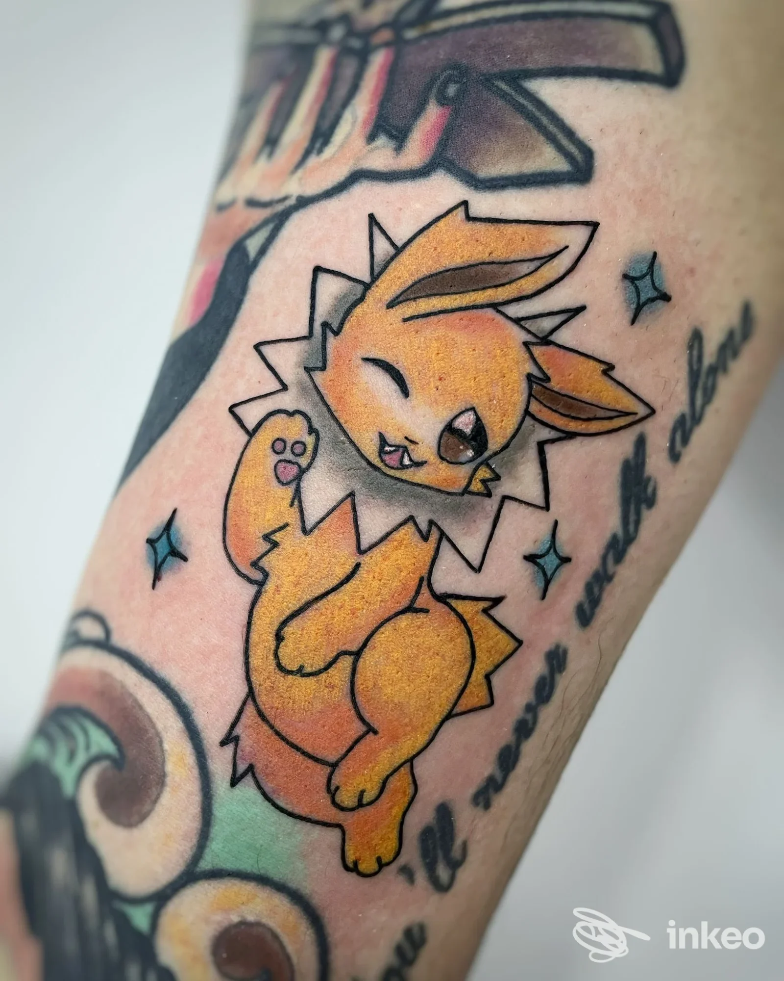 Tattoo colorato di un Pokémon sul braccio, stile cartone animato, contorni neri netti, colore giallo/arancio, sfondo pelle con dettagli circostanti della pelle.